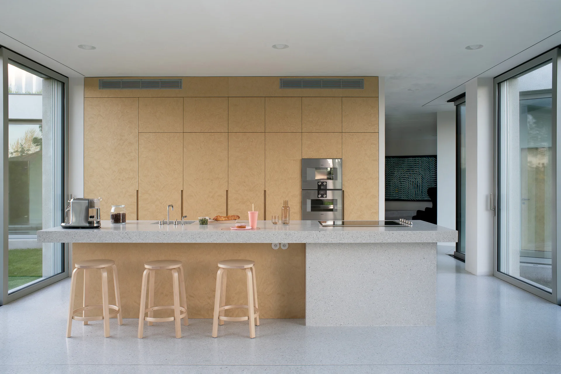 A minimalist kitchen features a large speckled stone island with three wooden stools in front of a full wall of light wood cabinetry.