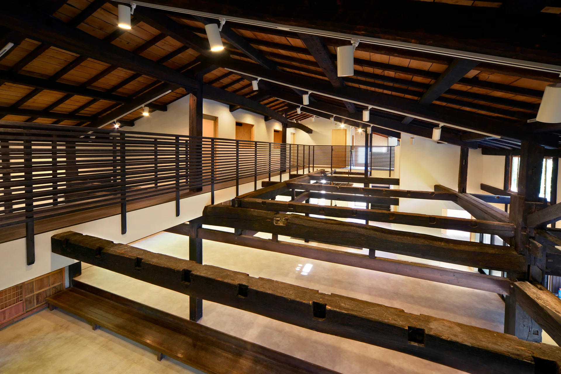 A modern mezzanine level with a dark slatted wood railing overlooks a spacious room with massive exposed timber beams and a wood plank ceiling.