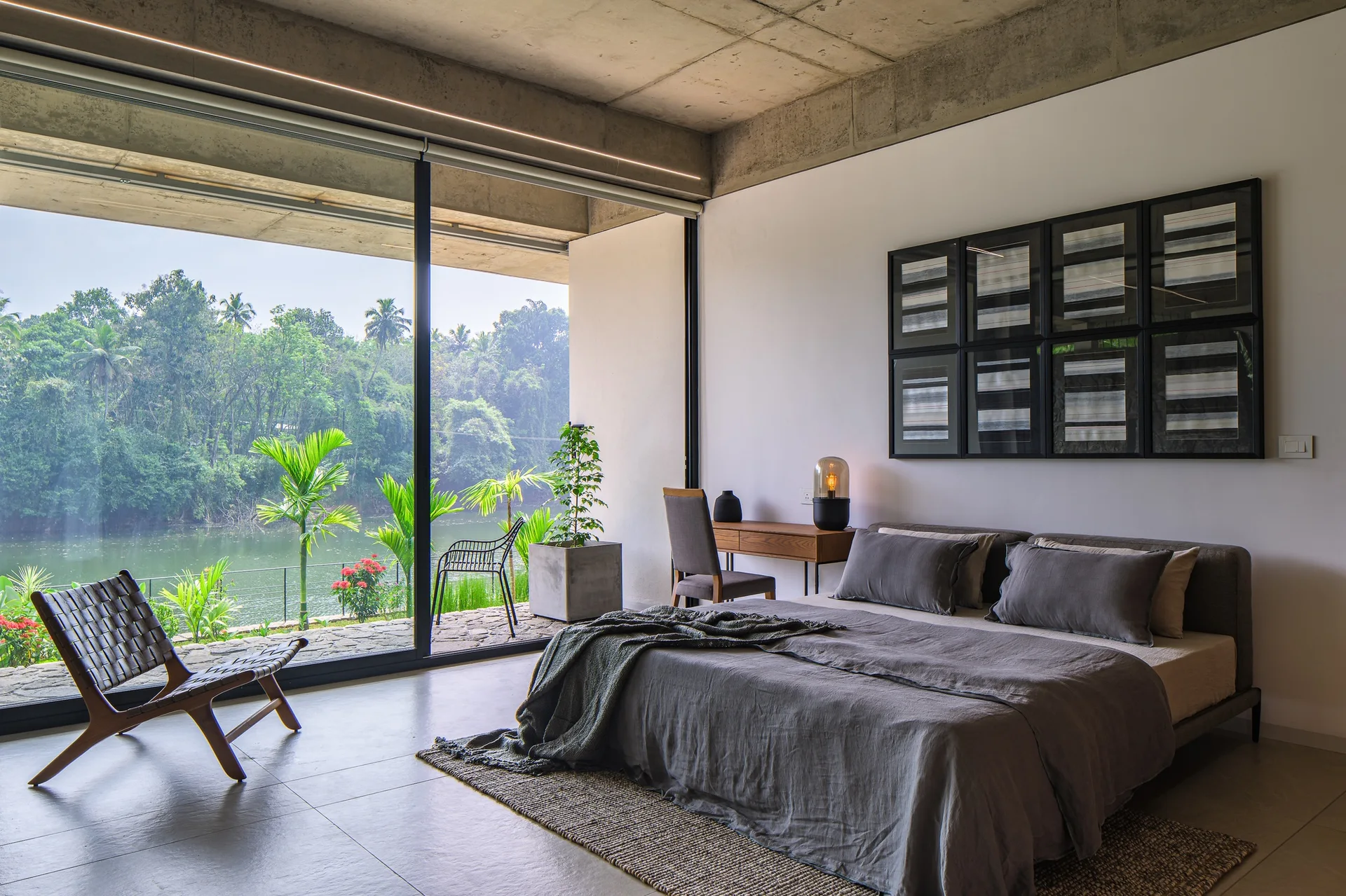 A modern bedroom features a grey low-profile bed, an exposed concrete ceiling, a woven leather lounge chair, and large sliding glass doors.