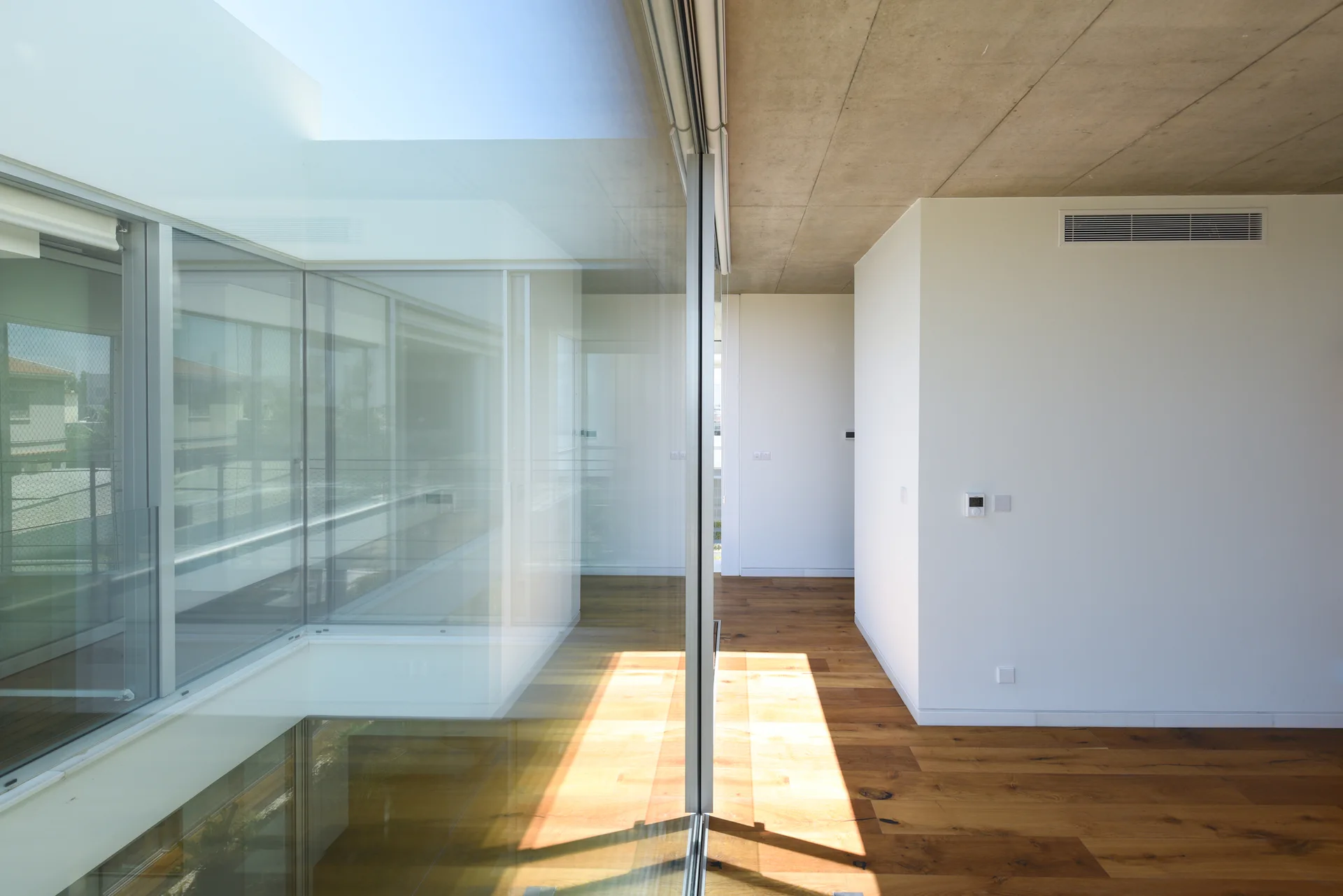 Minimalist interior hallway with floor-to-ceiling glass walls, an exposed concrete ceiling, and warm wood floors under bright natural light.