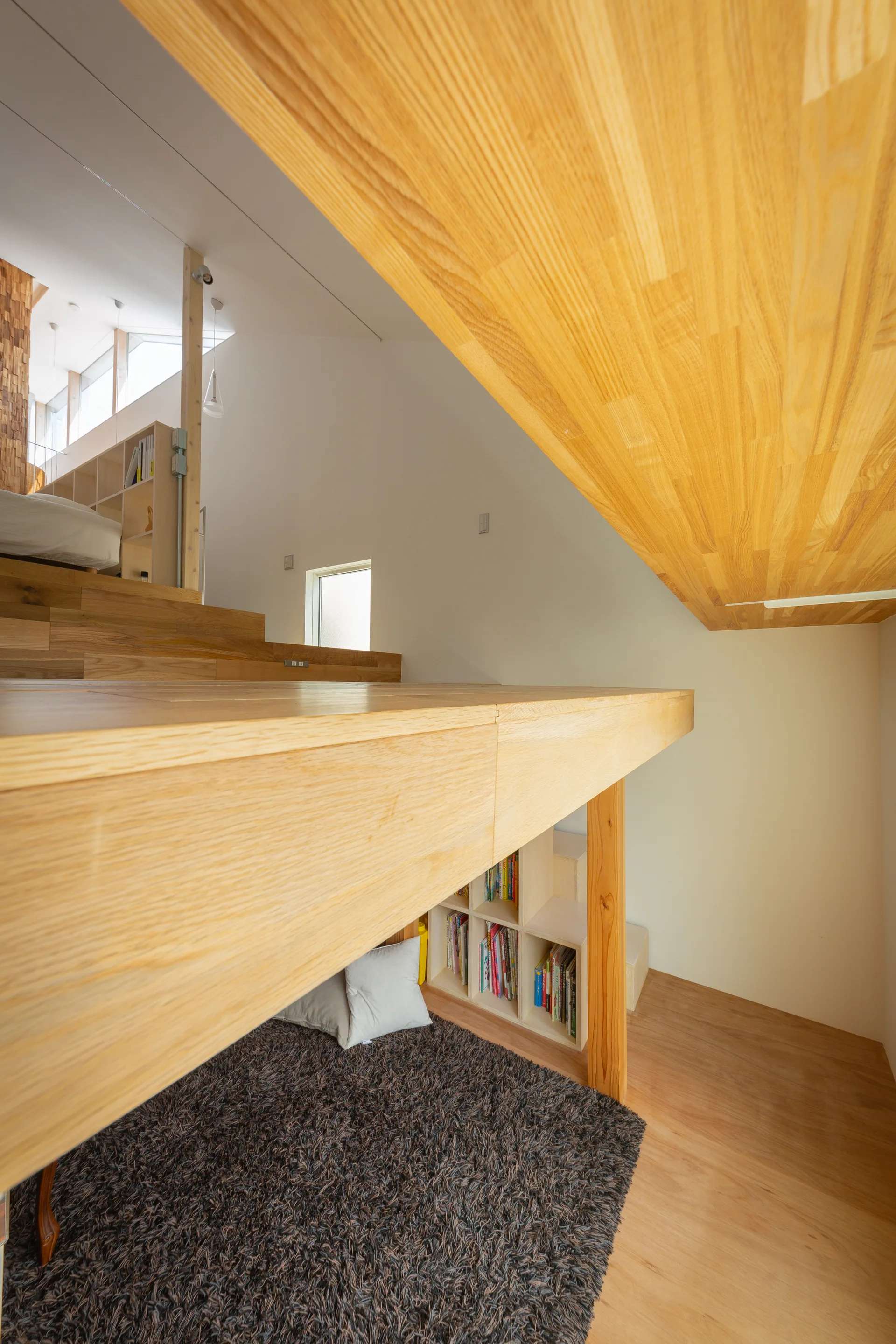 A split-level interior featuring a natural wood platform, a low cubby bookshelf tucked underneath, a dark shaggy rug, and white pillows.