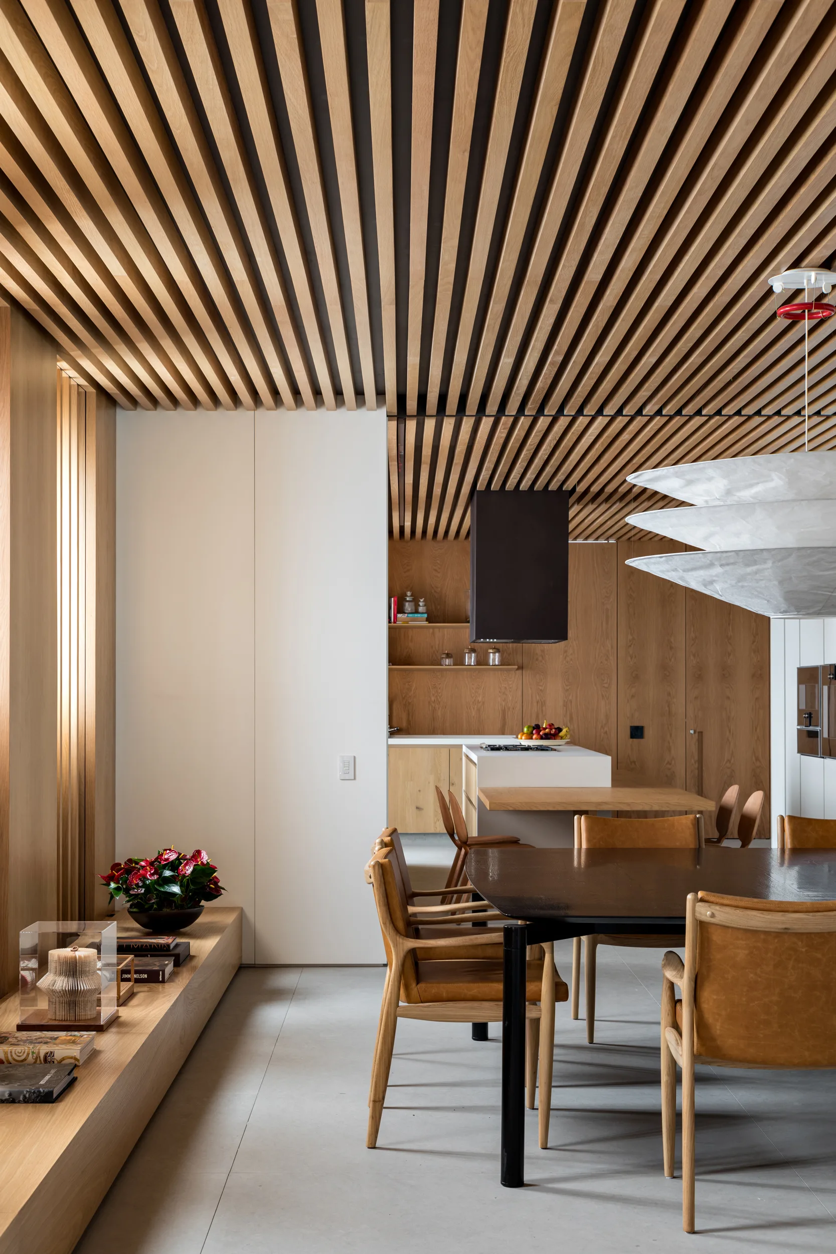 A modern dining room features a dark table and leather chairs beneath a slatted wood ceiling that extends into the open kitchen.