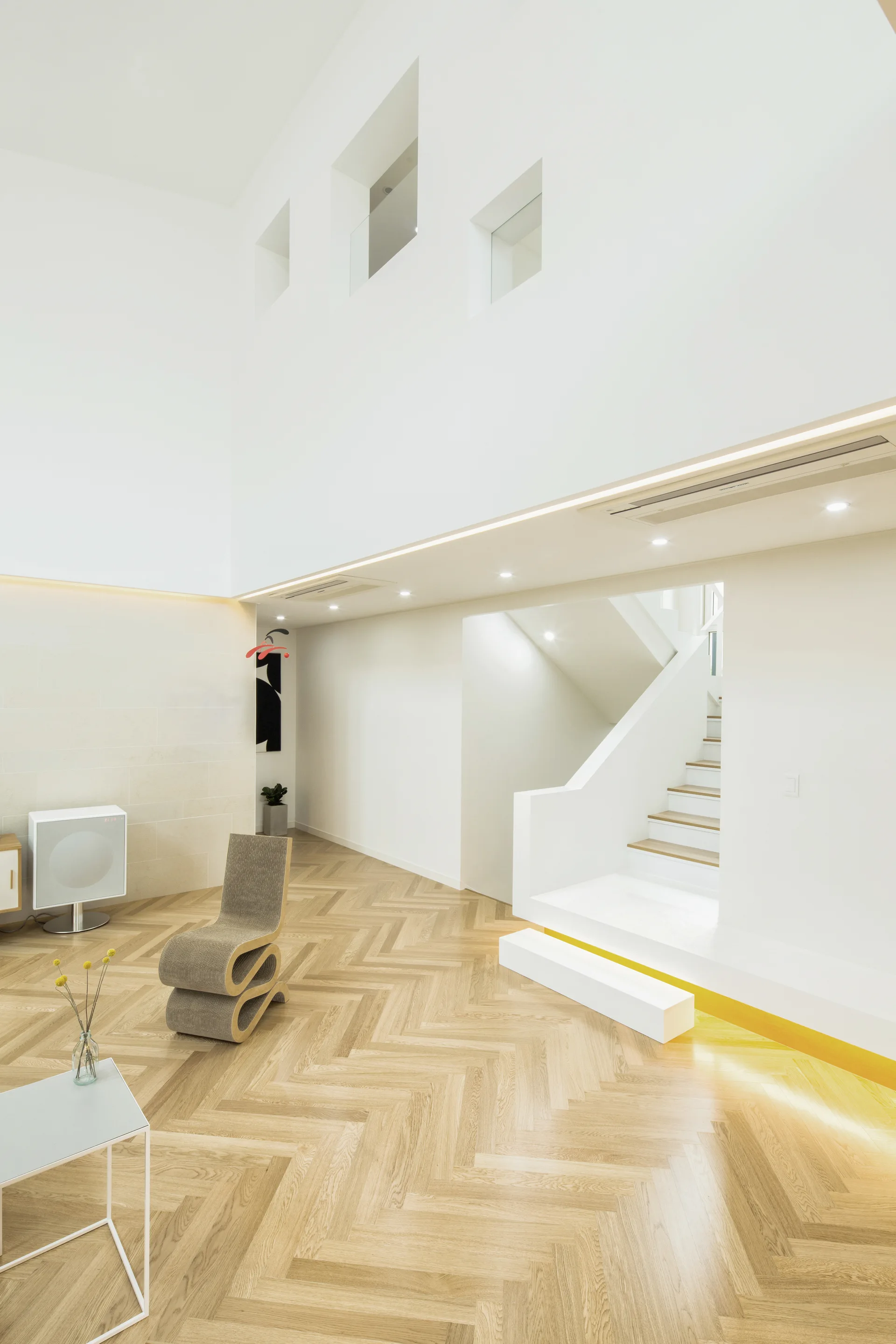 A bright, minimalist living room featuring light wood herringbone floors, a sculptural grey chair, and a white staircase under a double-height ceiling.