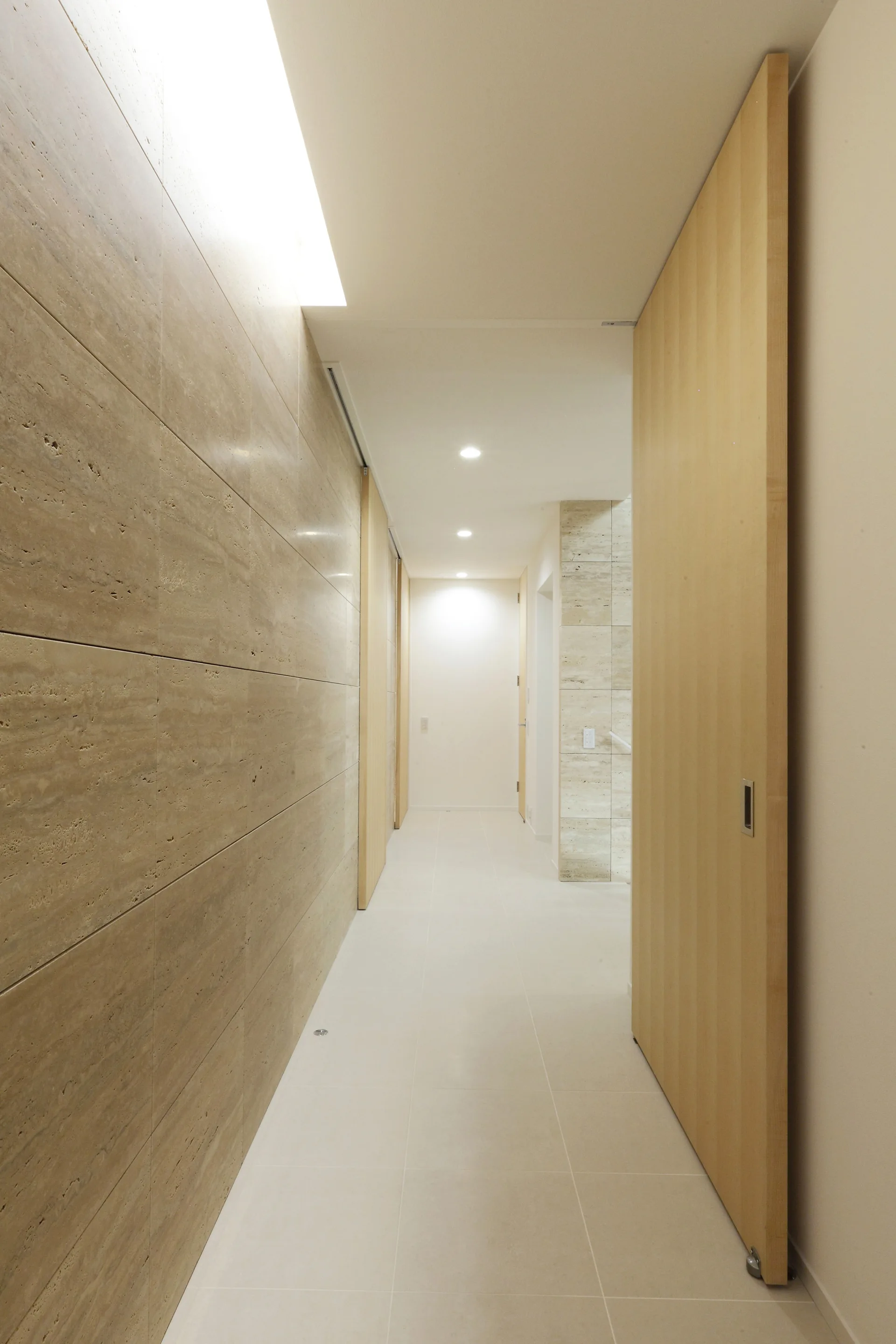 A minimalist residential hallway features a textured beige stone panel wall on the left, a tall light wood door on the right, and light floor tiles.