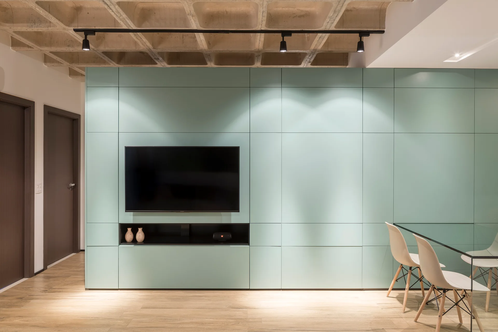 A modern interior features a mint green paneled wall with a television, a concrete waffle ceiling, and a glass dining table with white chairs.