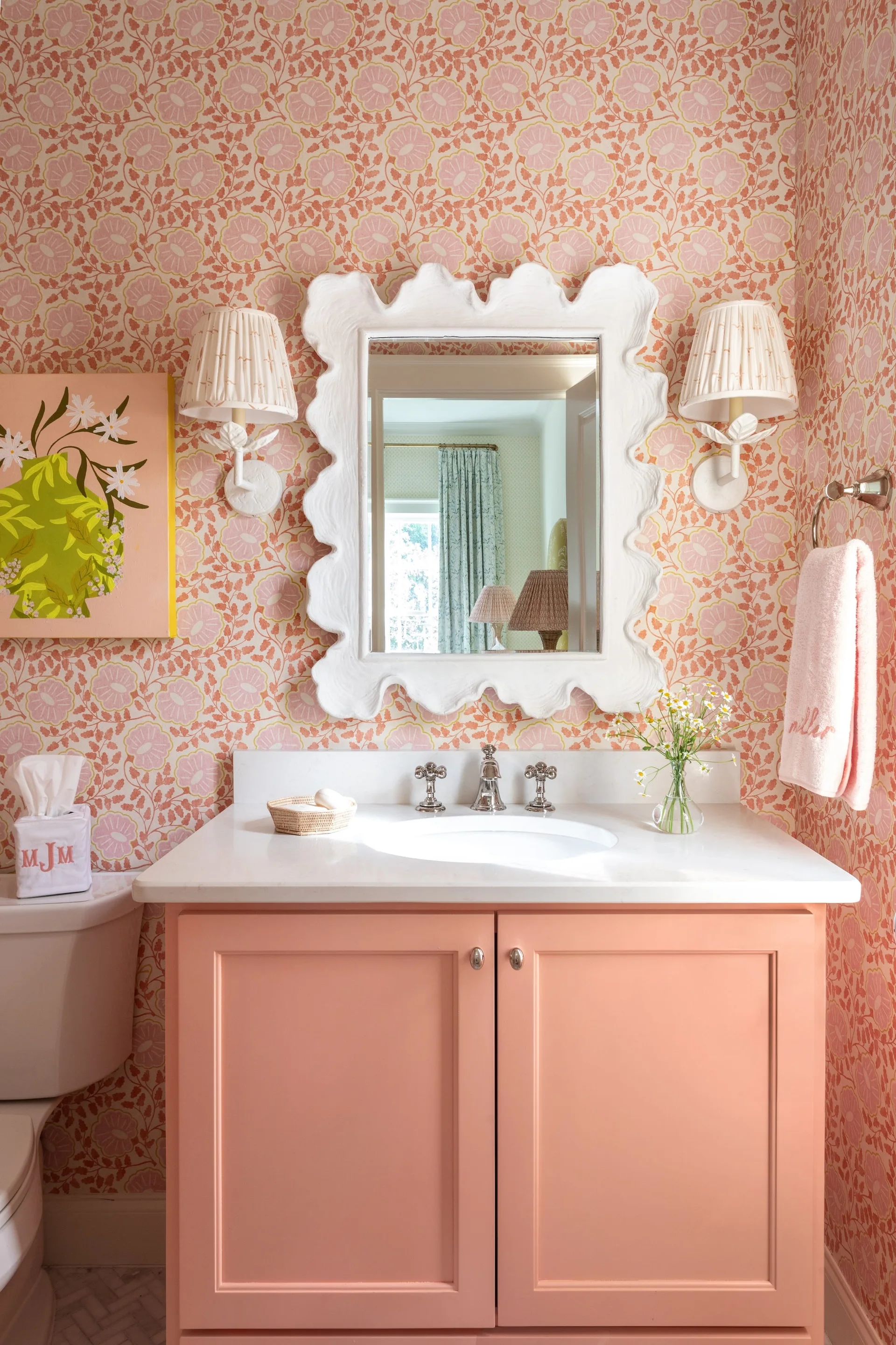 A vibrant powder room features a pink vanity cabinet, white scalloped-edge mirror, and walls covered in pink floral wallpaper.
