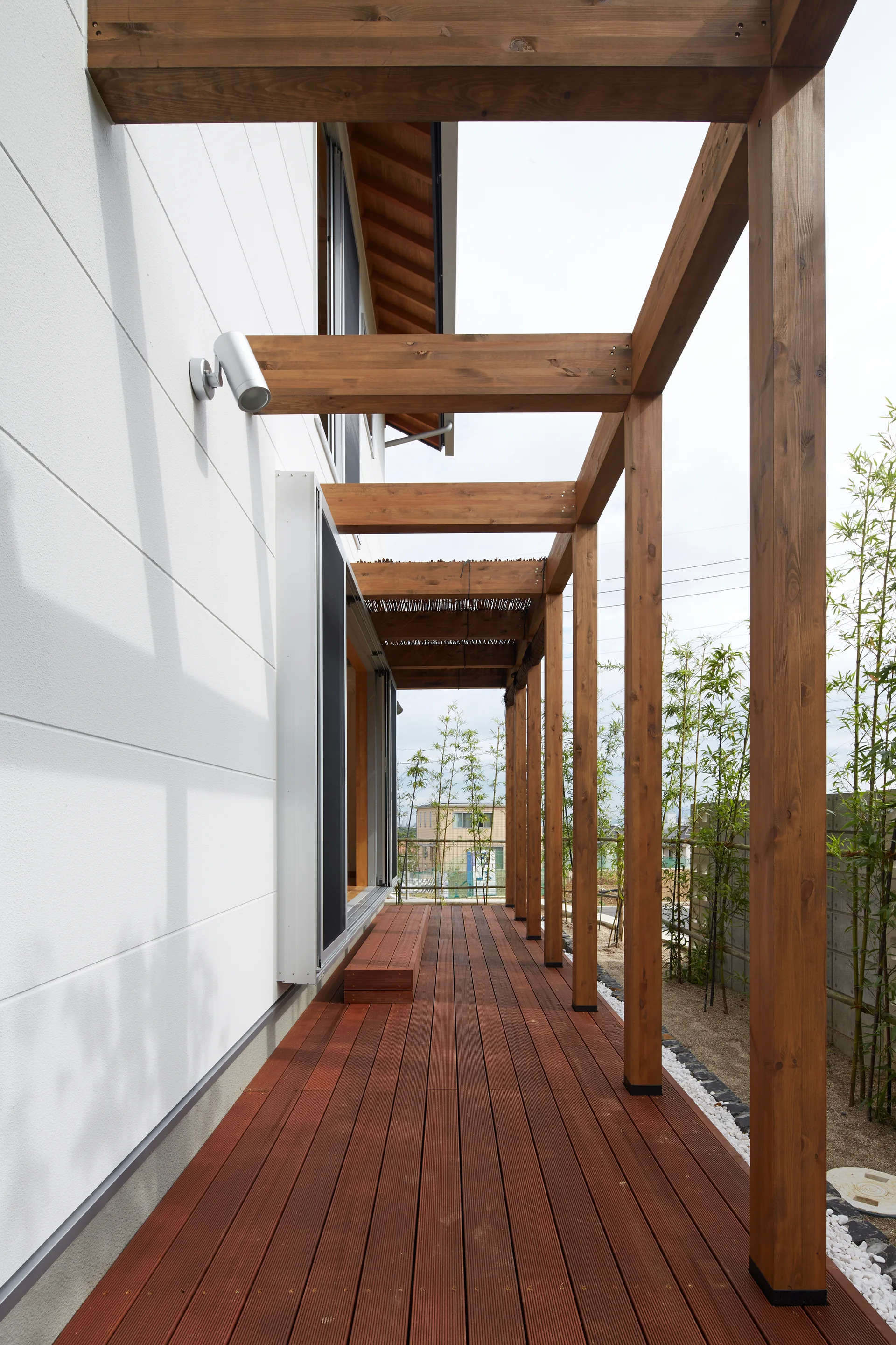 A long wooden deck with a matching pergola structure runs alongside a white modern house next to green bamboo plants.