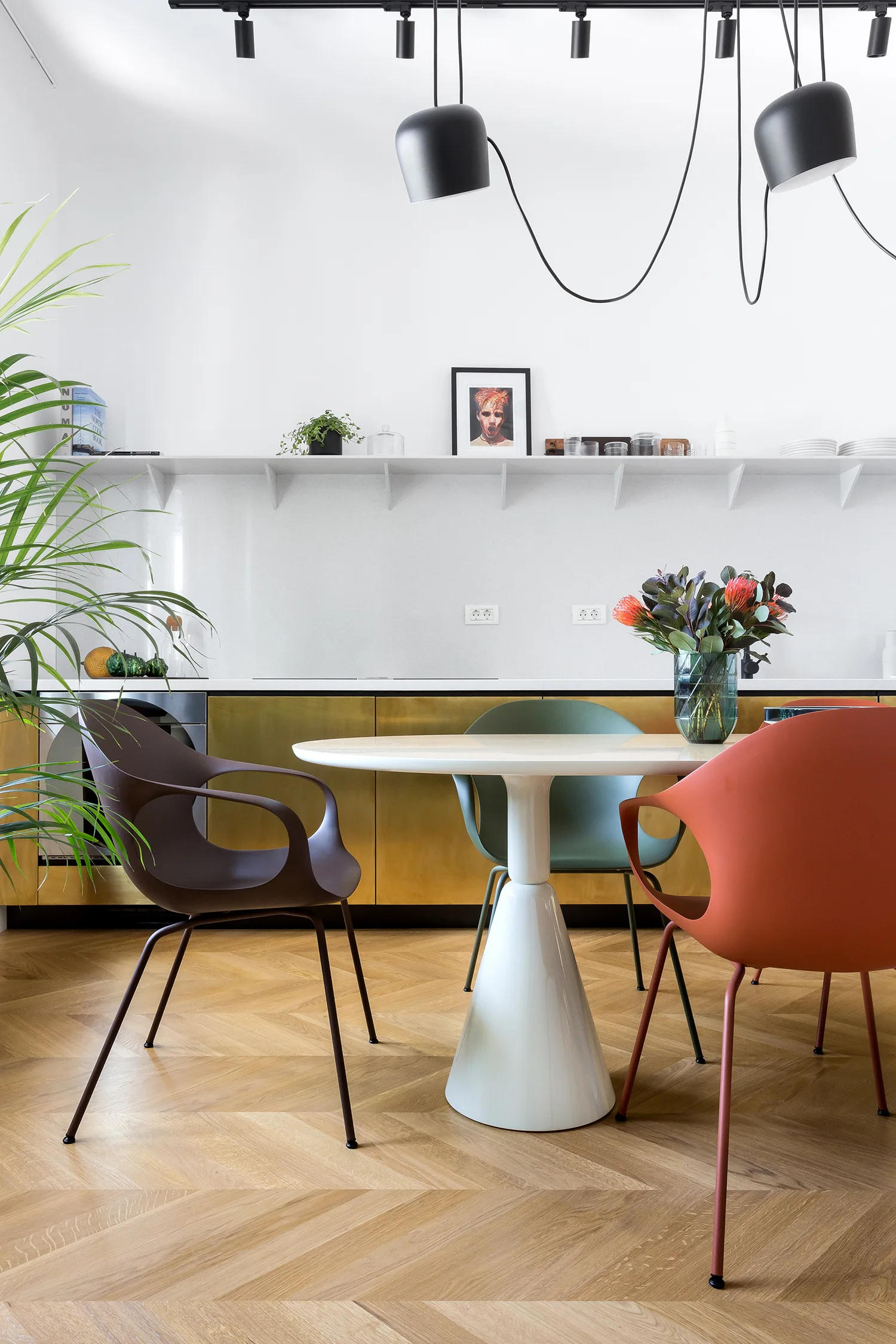 A modern kitchen dining area features a white tulip table with colorful molded chairs, metallic gold cabinets, and light wood herringbone flooring.