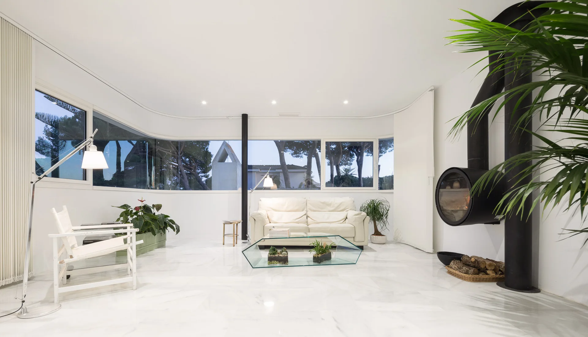 A minimalist white living room features a curved glass window wall, white marble floors, a leather sofa, and a sculptural black fireplace.