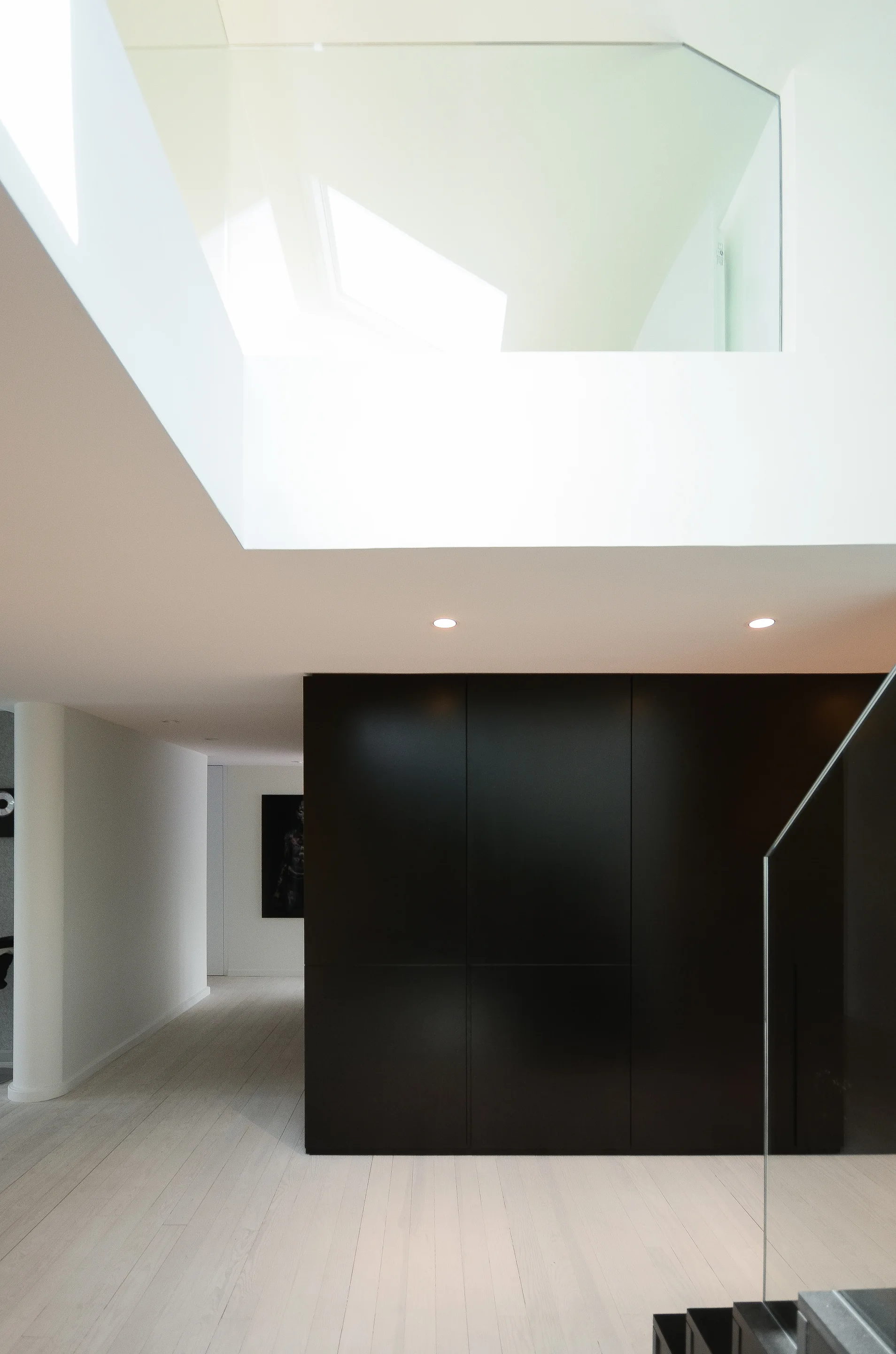 A minimalist hallway featuring light wood floors, a large matte black storage cabinet, and a staircase with a transparent glass railing under a skylight.