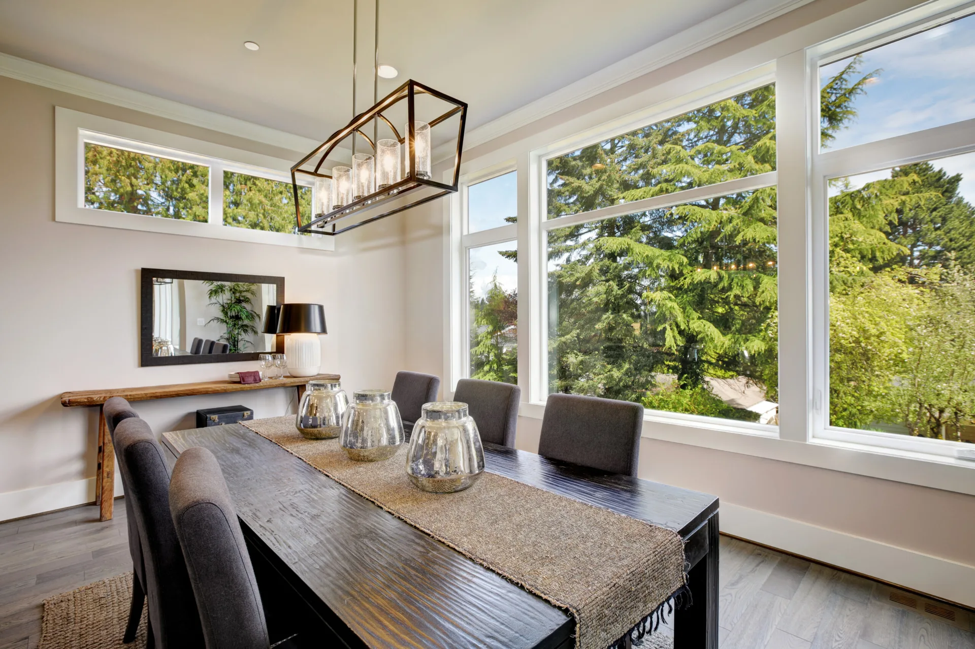 A modern dining room featuring a dark wood table with grey upholstered chairs, a rectangular metal chandelier, and large windows looking out to trees.