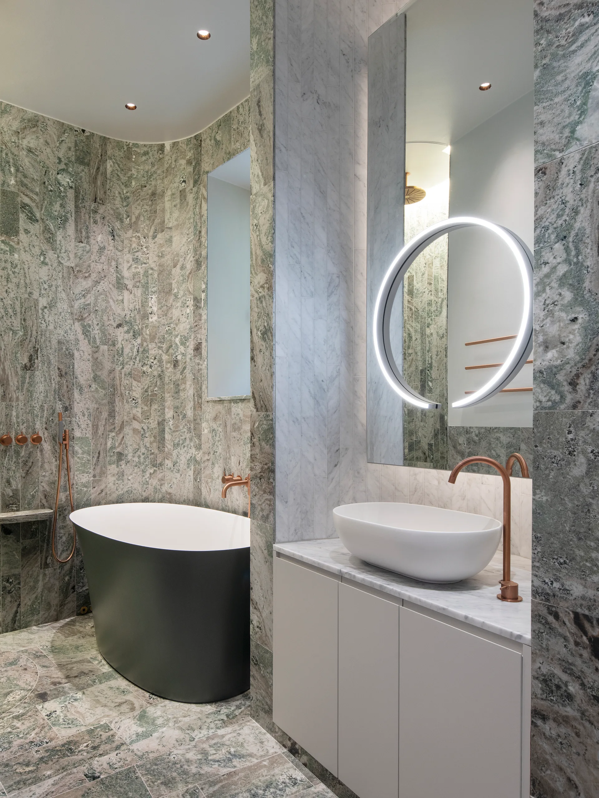 A modern bathroom featuring green marble walls, a matte black freestanding bathtub, and a white vanity with a vessel sink and copper fixtures.
