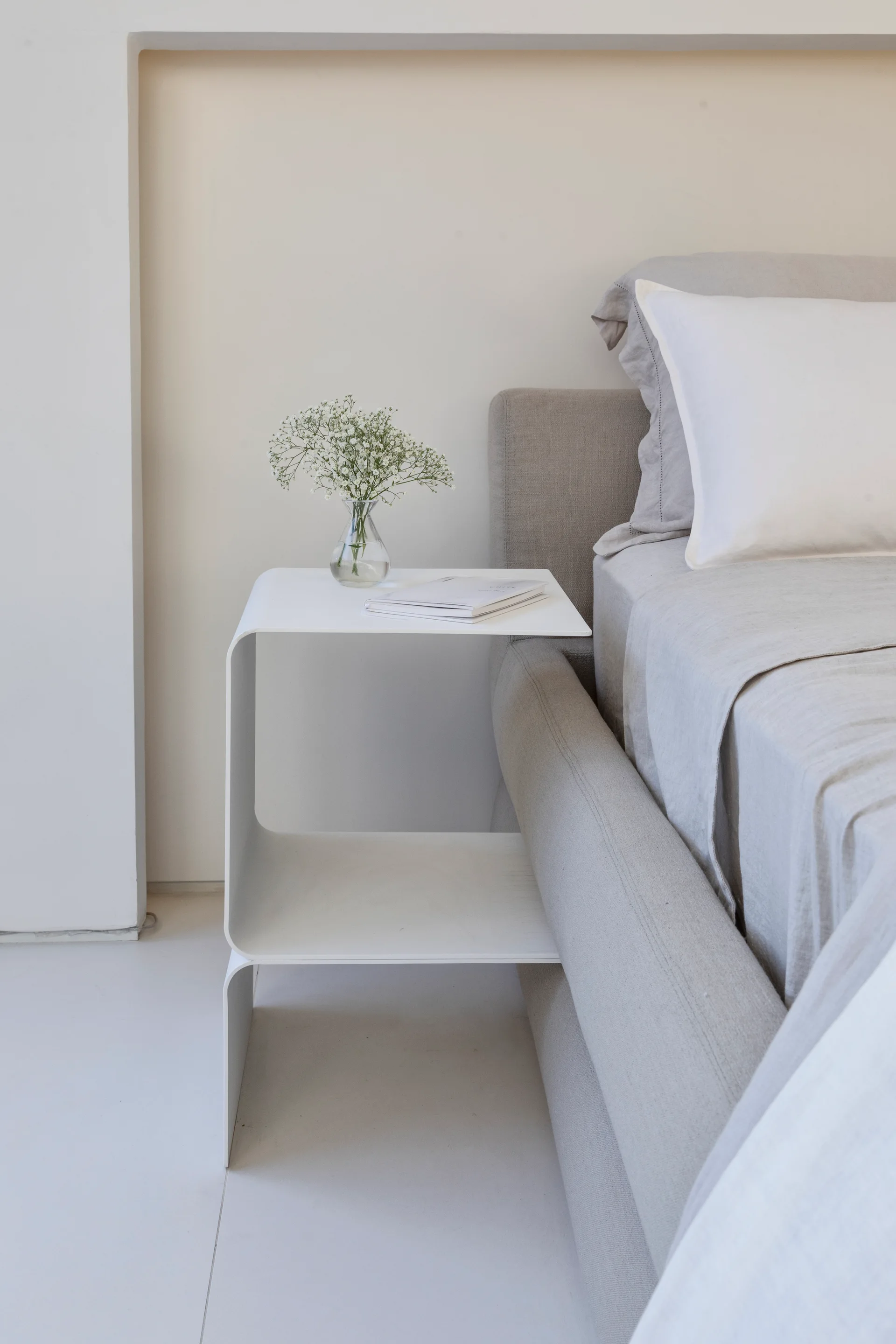 A light grey upholstered bed sits beside a minimalist white metal C-shaped nightstand styled with a small glass vase of flowers and a book.