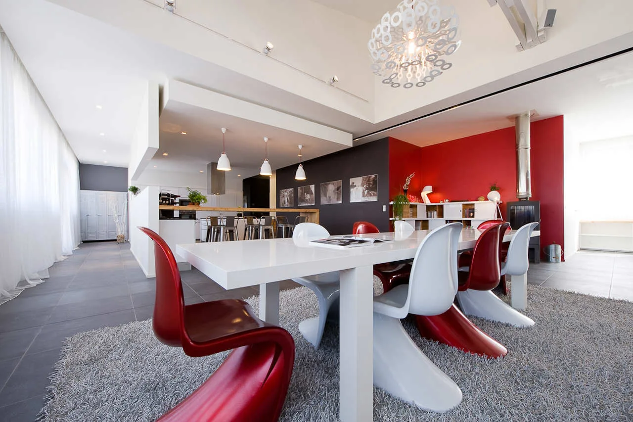 An open-concept dining room features a white table with glossy red and white cantilever chairs atop a grey shag rug.