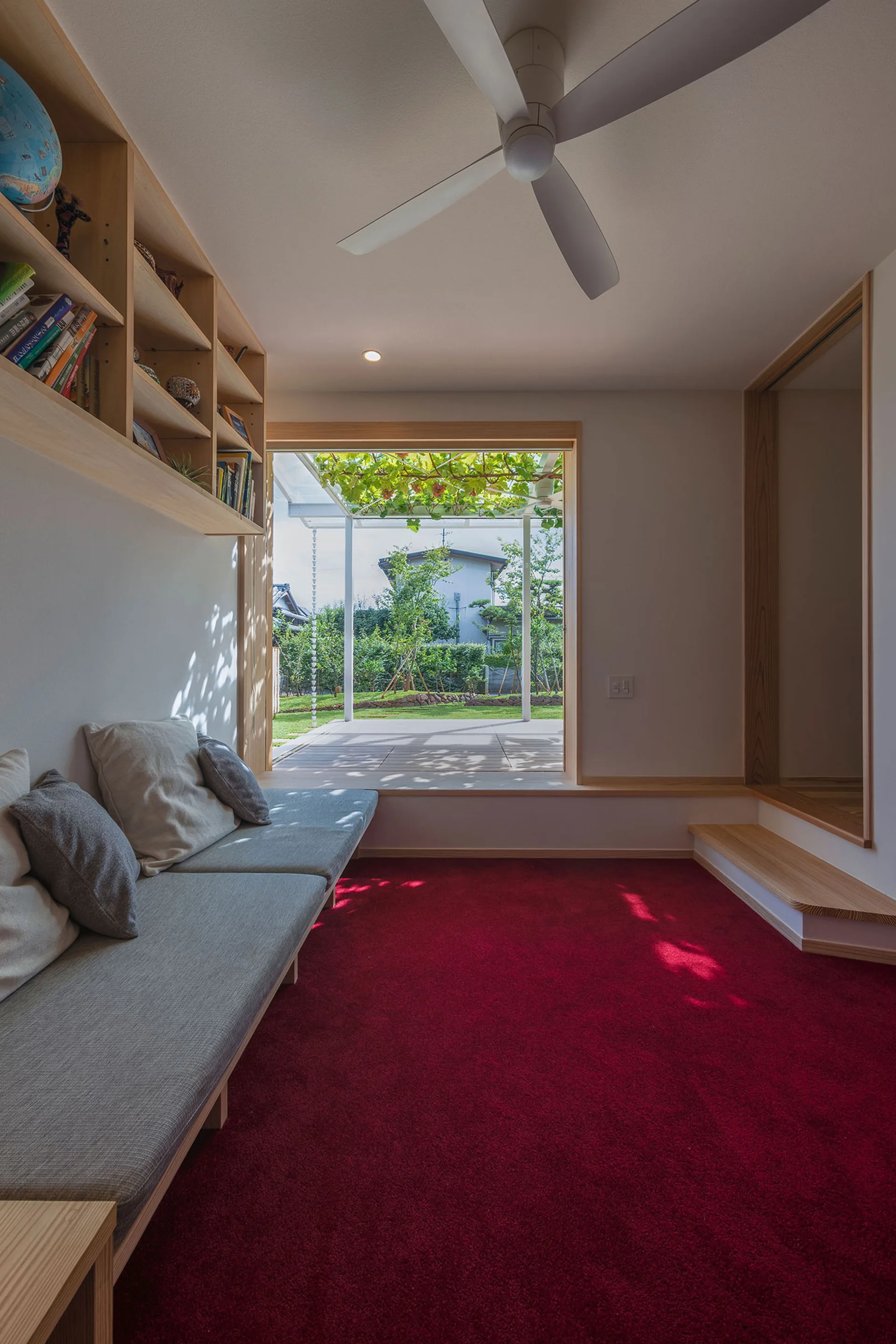 A minimalist room features a bright red carpeted floor, a long built-in wooden daybed with grey pillows, and a large window framing a garden.