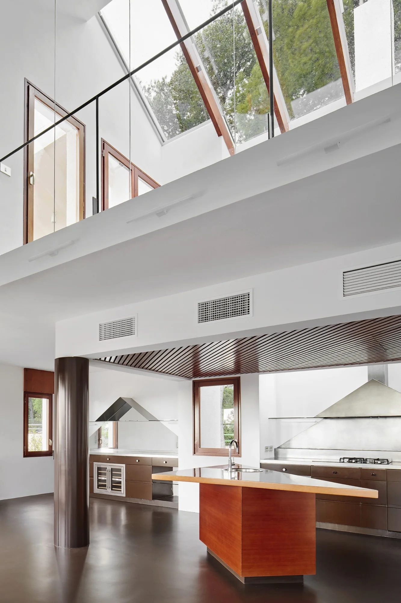 A modern kitchen features a wood-fronted island, a slatted ceiling detail, and dark floors beneath a large sloped glass skylight.