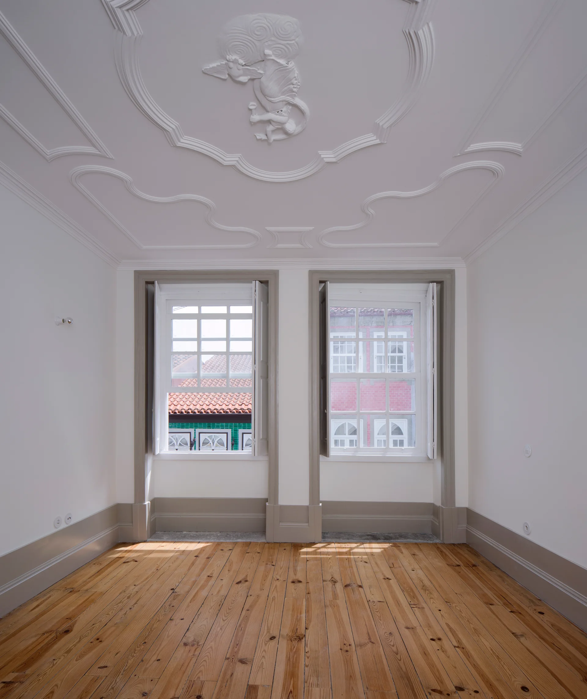 An empty room with an ornate white plaster ceiling molding, light wood floors, and two symmetrical windows framed in warm grey trim.