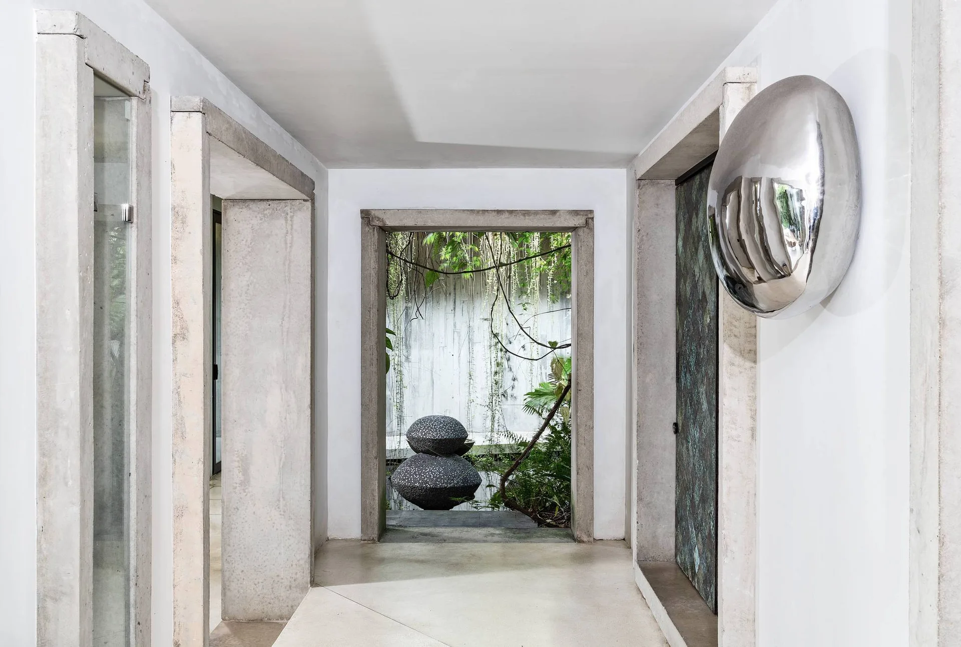 A white hallway with raw concrete frames features a reflective silver wall sculpture and a doorway opening to an outdoor garden with a stone sculpture.
