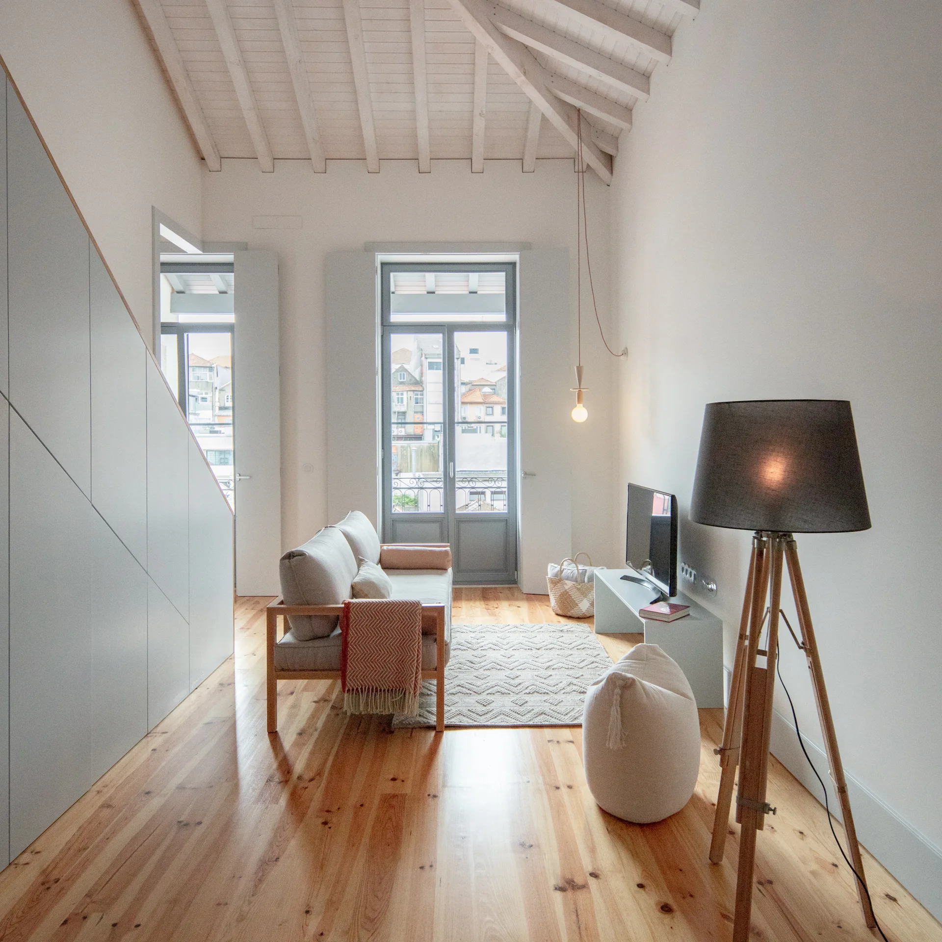 A narrow attic living room features a grey sofa, wood floors, a tripod lamp, and a tall balcony door under a vaulted beamed ceiling.