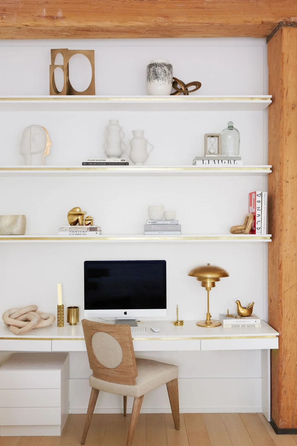 A white built-in desk nook with a computer, brass lamp, and upholstered wood chair below open shelves detailed with gold trim and neutral decor.