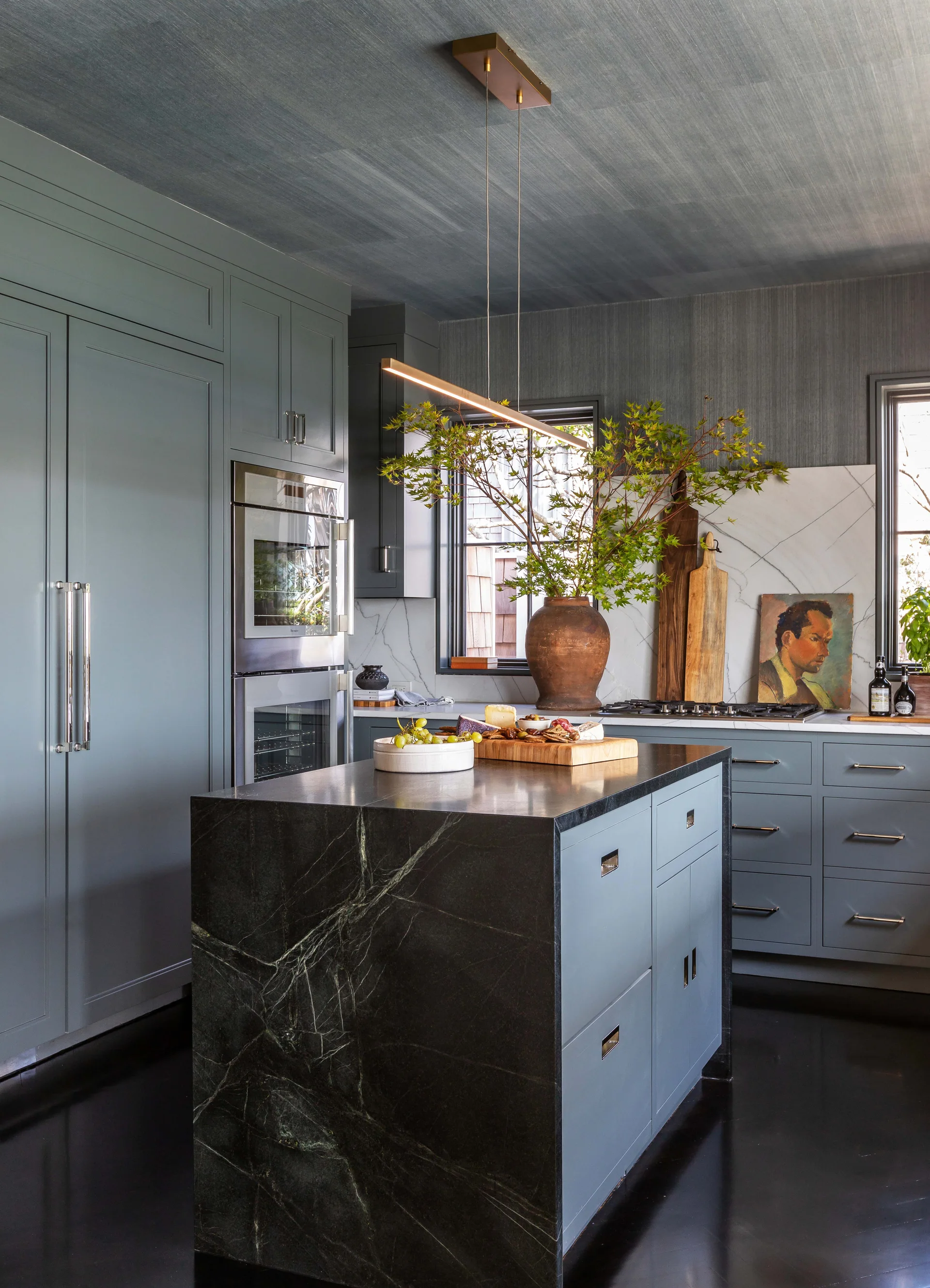 A blue-gray kitchen features a dark veined marble island, matching painted cabinets, a white slab backsplash, and a textured grasscloth ceiling.