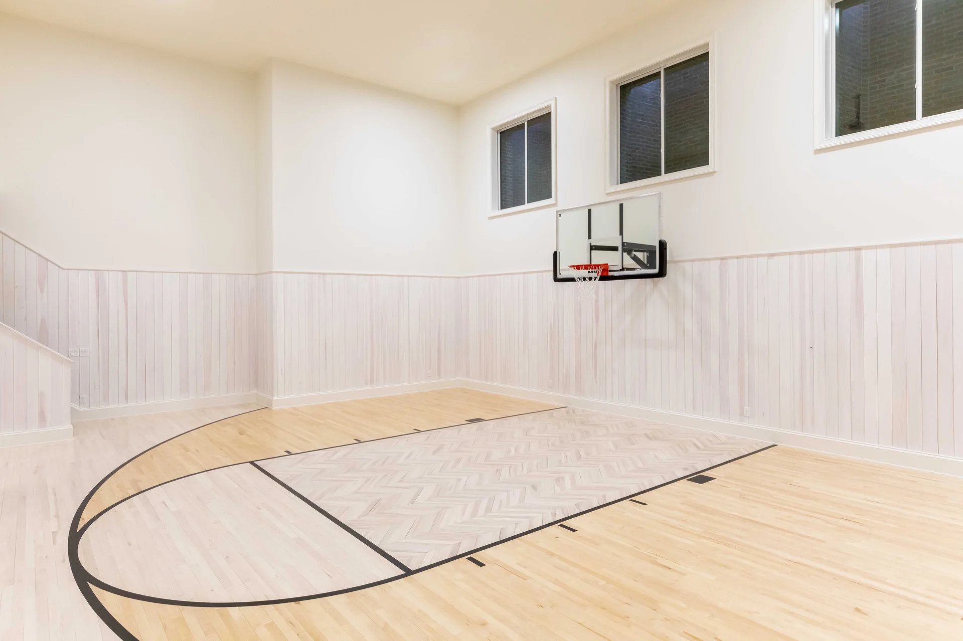 An indoor basketball court features light hardwood flooring with a herringbone-patterned key, whitewashed vertical wainscoting, and a wall-mounted hoop.