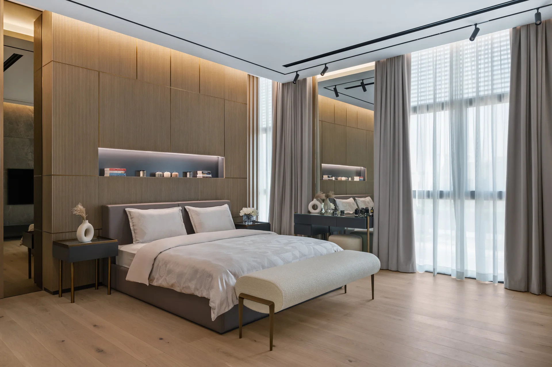 A modern bedroom features a bed with beige bedding set against a slatted wood paneled wall with a lit recessed shelf and long gray curtains.