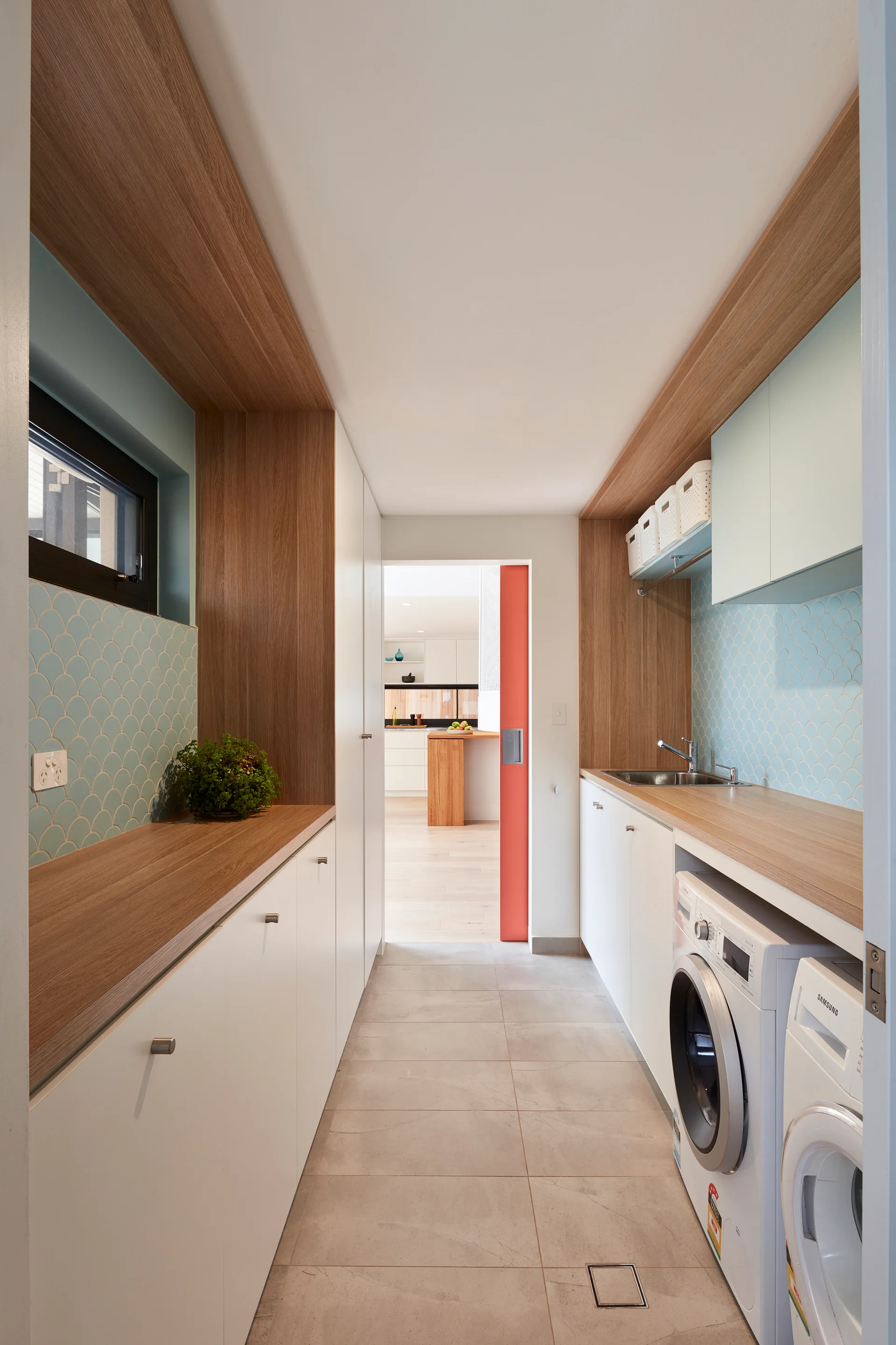A narrow modern laundry room features white cabinets, wood countertops, light blue fish-scale tile backsplashes, and a vibrant orange sliding door.