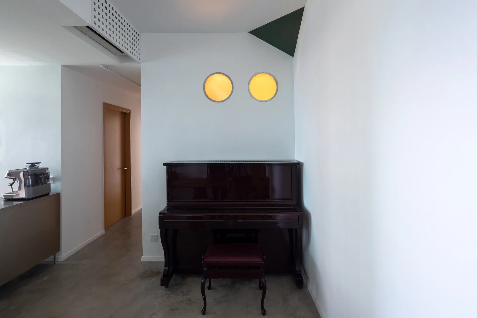 A dark wood upright piano stands on a concrete floor beneath two glowing yellow circular wall lights in a minimalist white interior.