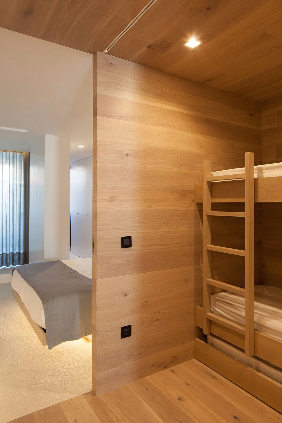 A bedroom split by a light wood wall features a minimalist bed with under-bed lighting next to an integrated wooden bunk bed nook.
