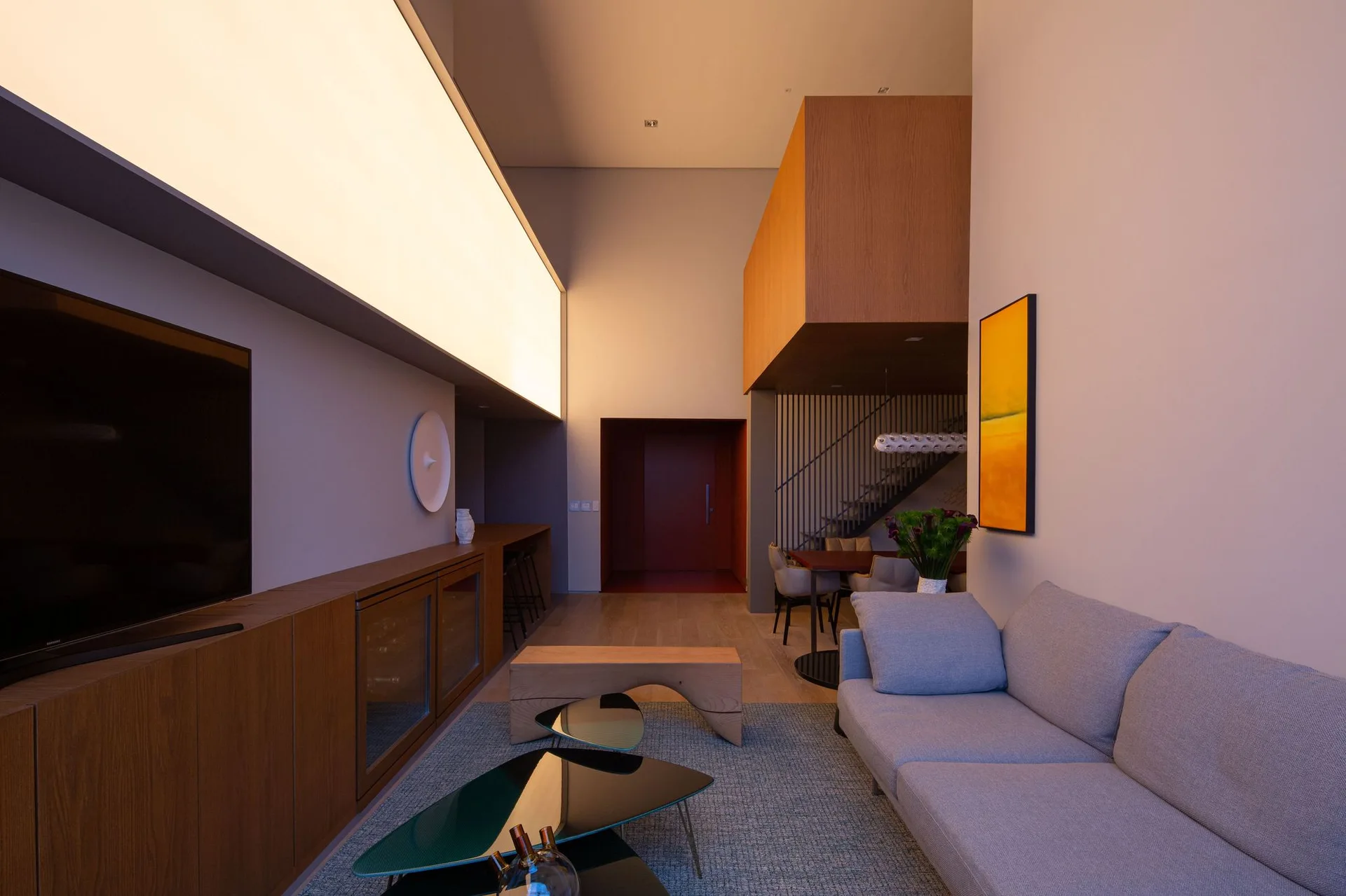 A modern double-height living room featuring a grey sofa, nested black glass coffee tables, a long wooden console, and a large illuminated ceiling light.