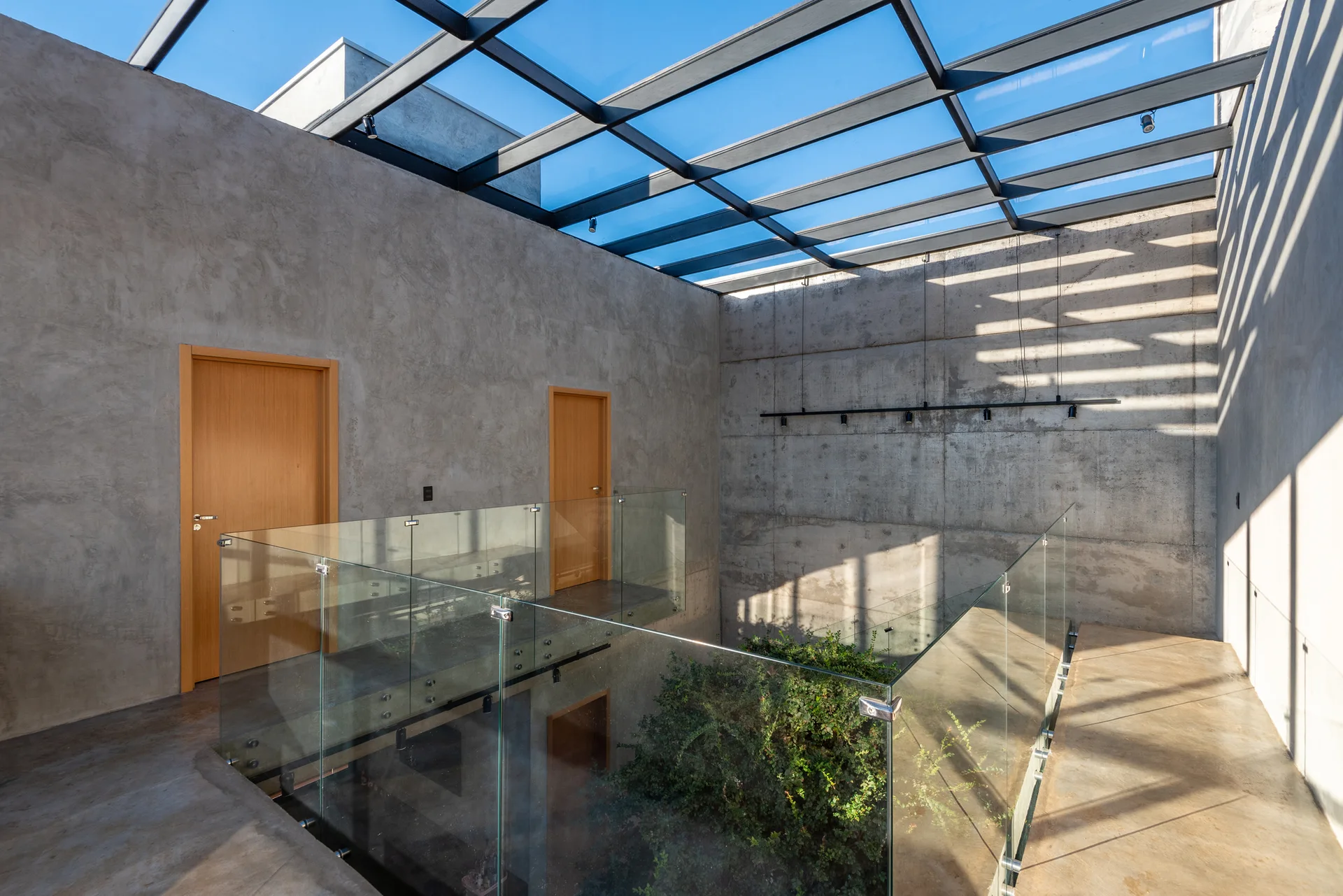 A concrete residential landing features frameless glass railings under a black metal grid skylight roof with wooden doors.