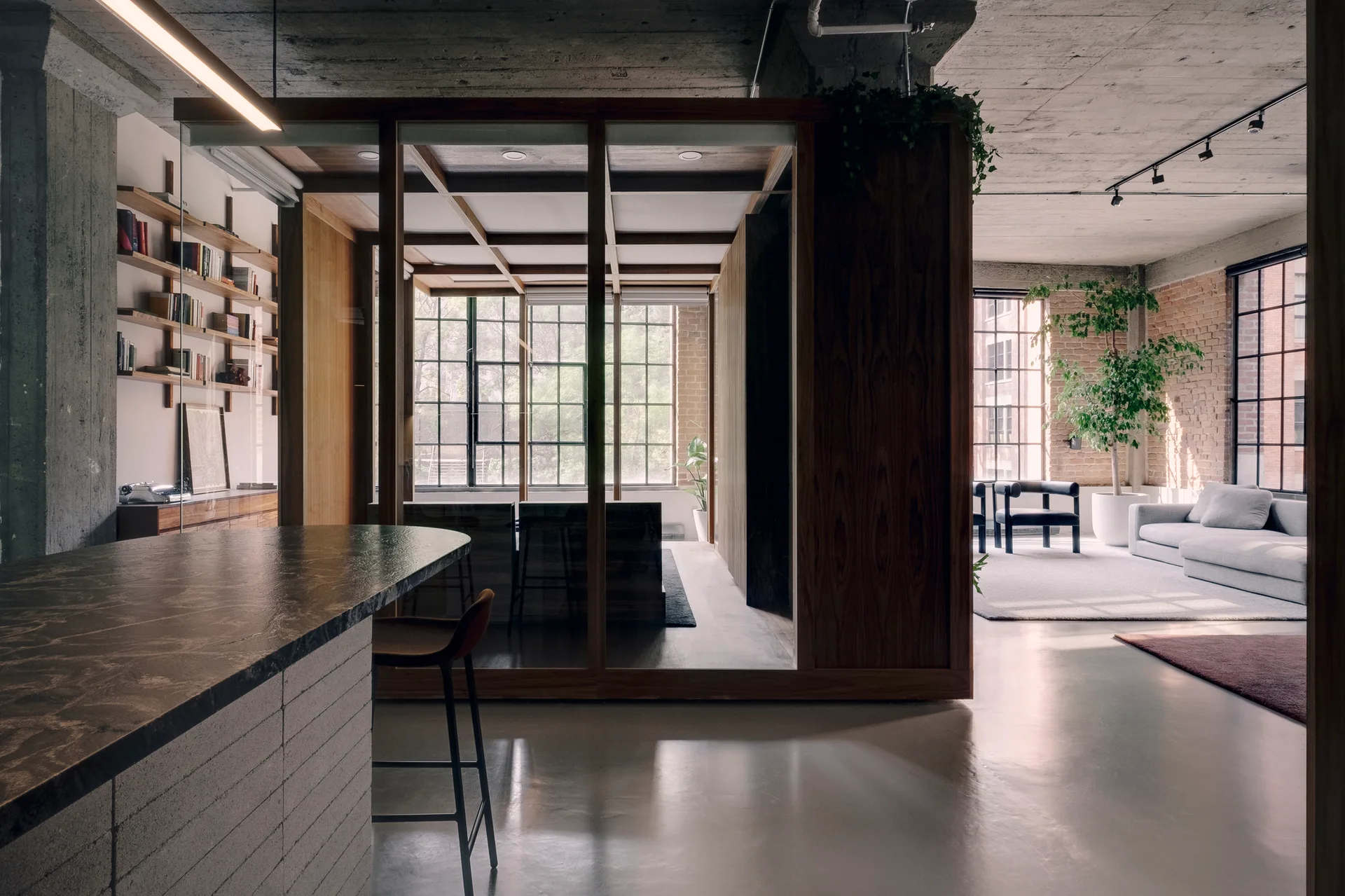 An industrial loft space featuring a central wood and glass room partition, a stone kitchen island, and a distant living area with a grey sofa.