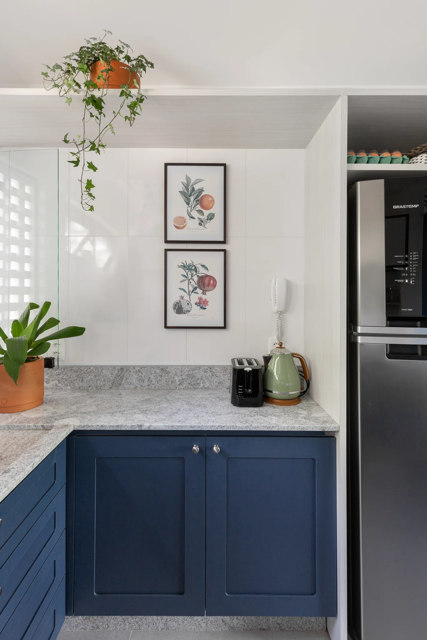 A kitchen corner features navy blue shaker cabinets, a speckled grey countertop, white tiled backsplash, and two stacked botanical prints.