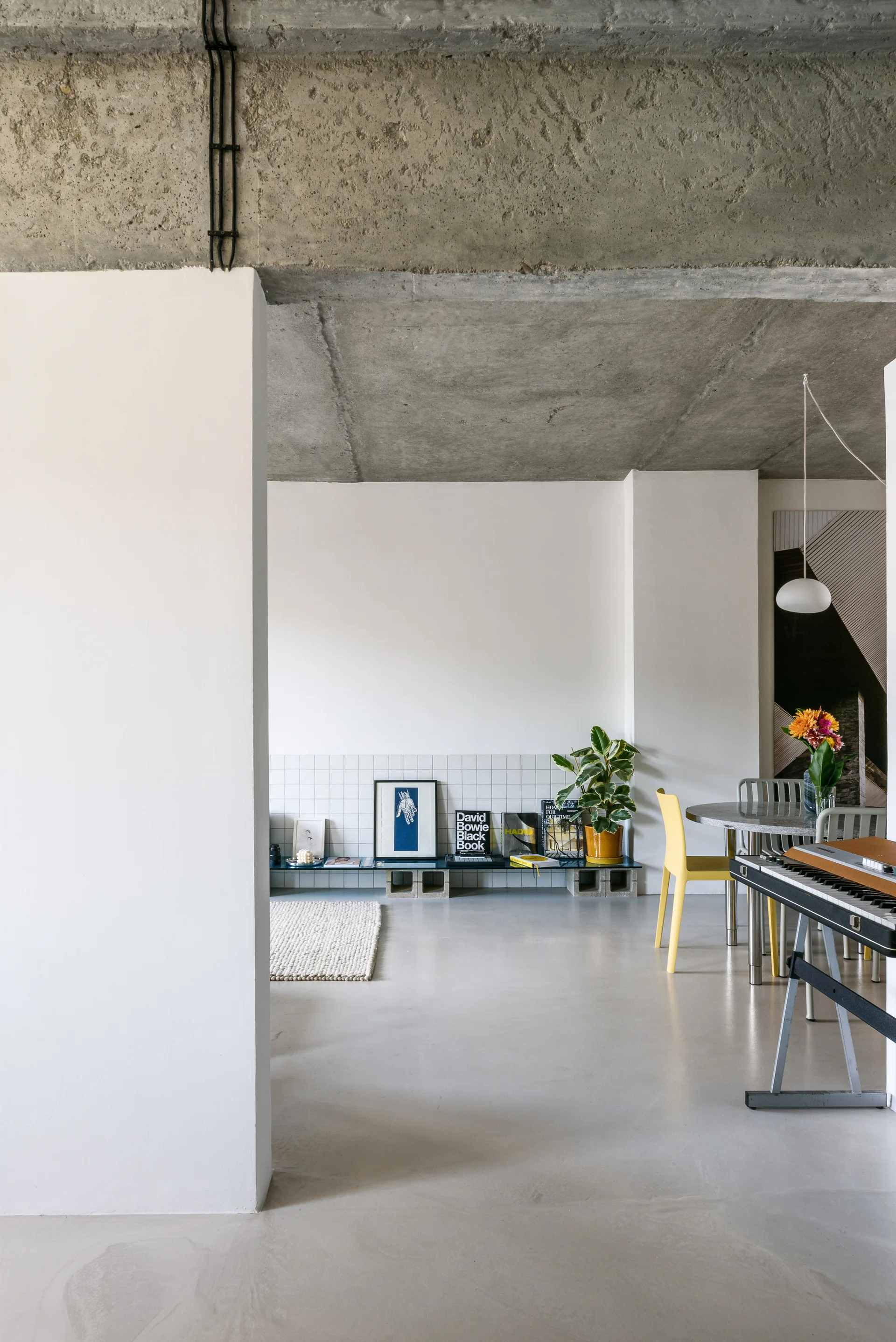 A modern minimalist living space features an exposed concrete ceiling, a white tiled lower wall, and a low display shelf resting on concrete blocks.