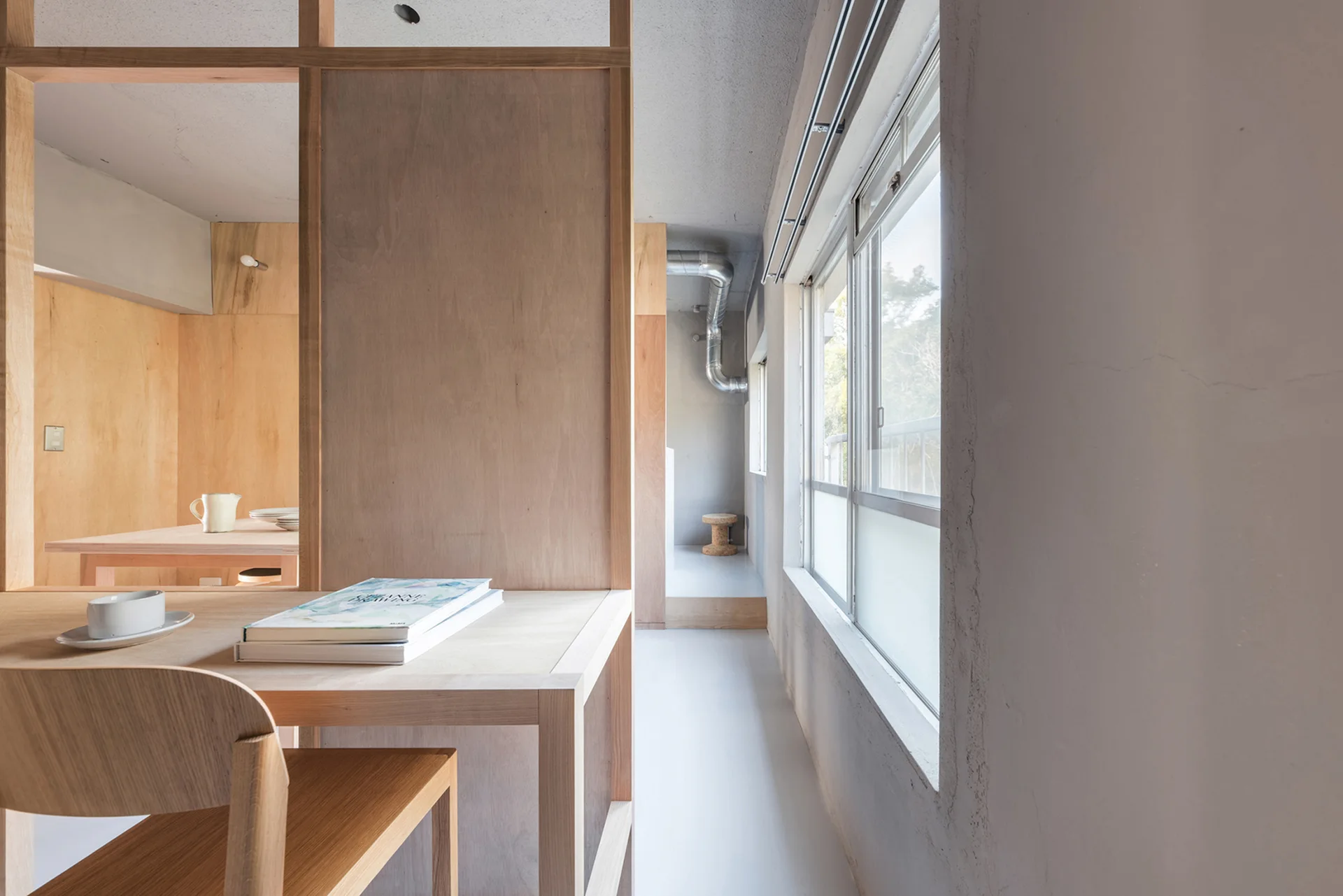 A minimalist interior features a light wood desk and chair set against a tall plywood partition wall, next to a concrete wall with windows.