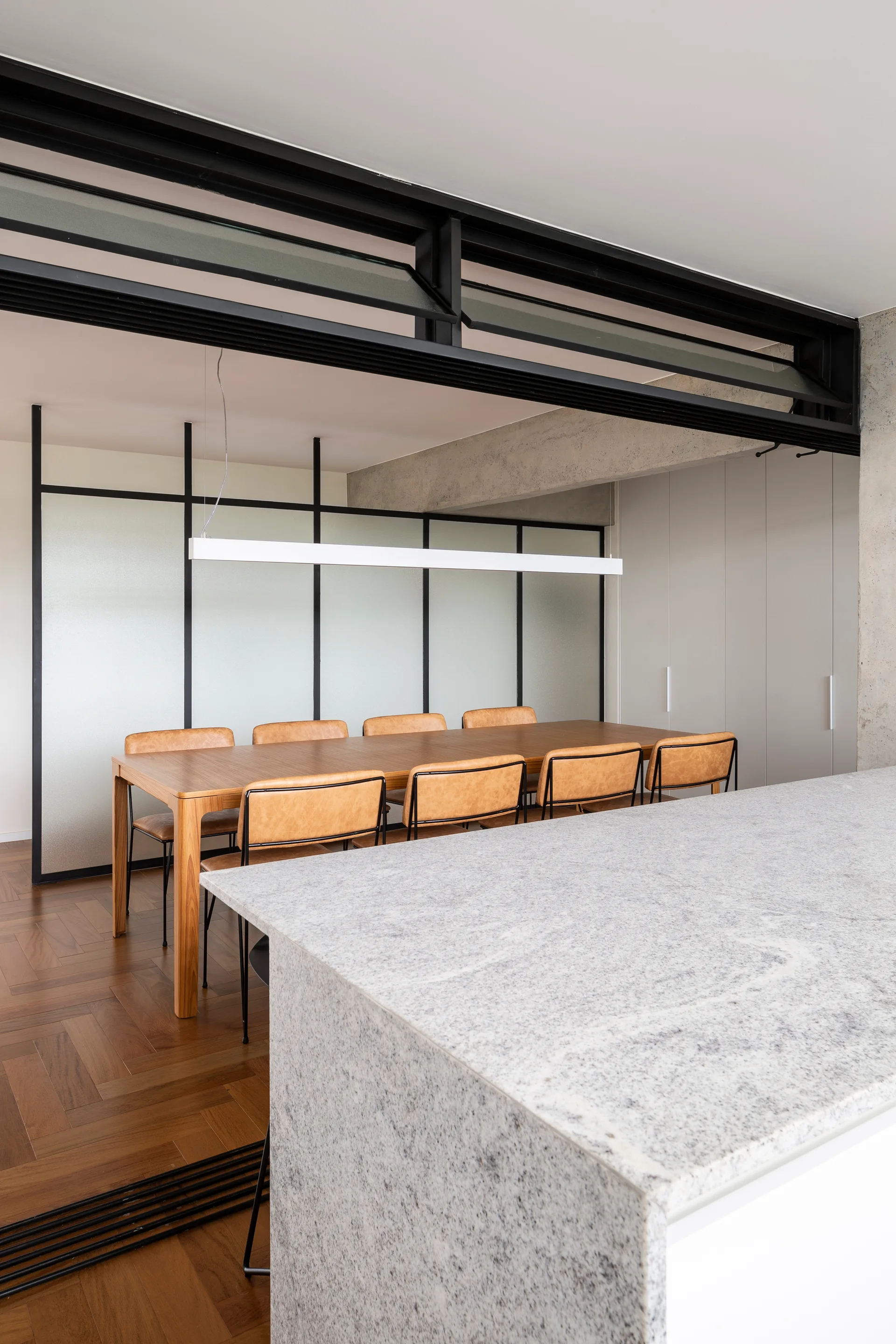A modern dining room with a wooden table, tan chairs, a black-framed frosted glass divider, and a stone-topped kitchen island in the foreground.