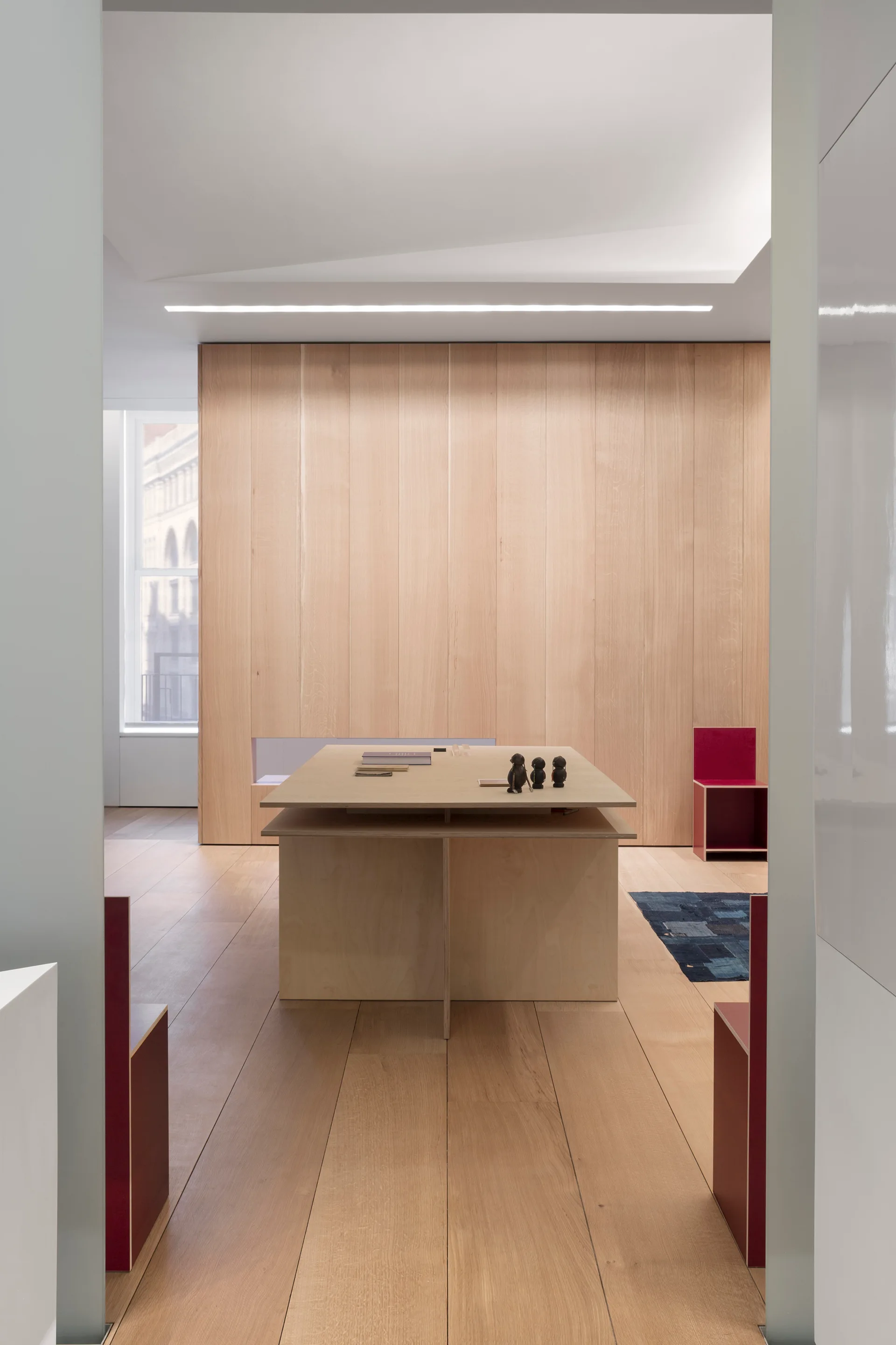 A minimalist studio workspace featuring a large central wood table, matching light wood wall paneling, and geometric red chairs.
