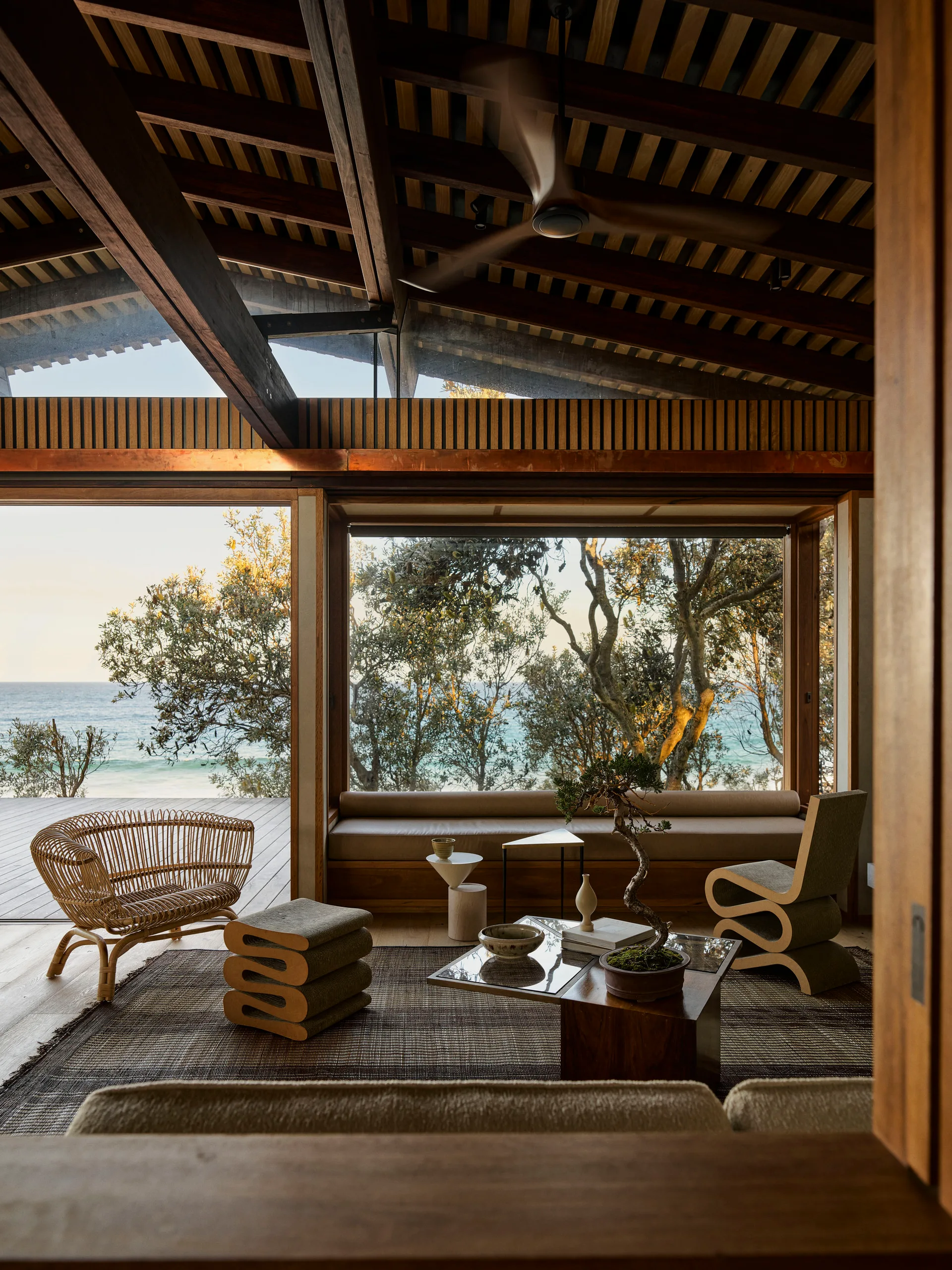 A coastal living room featuring a sloped wood ceiling, exposed timber beams, sculptural furniture, and a large window looking out at the ocean.