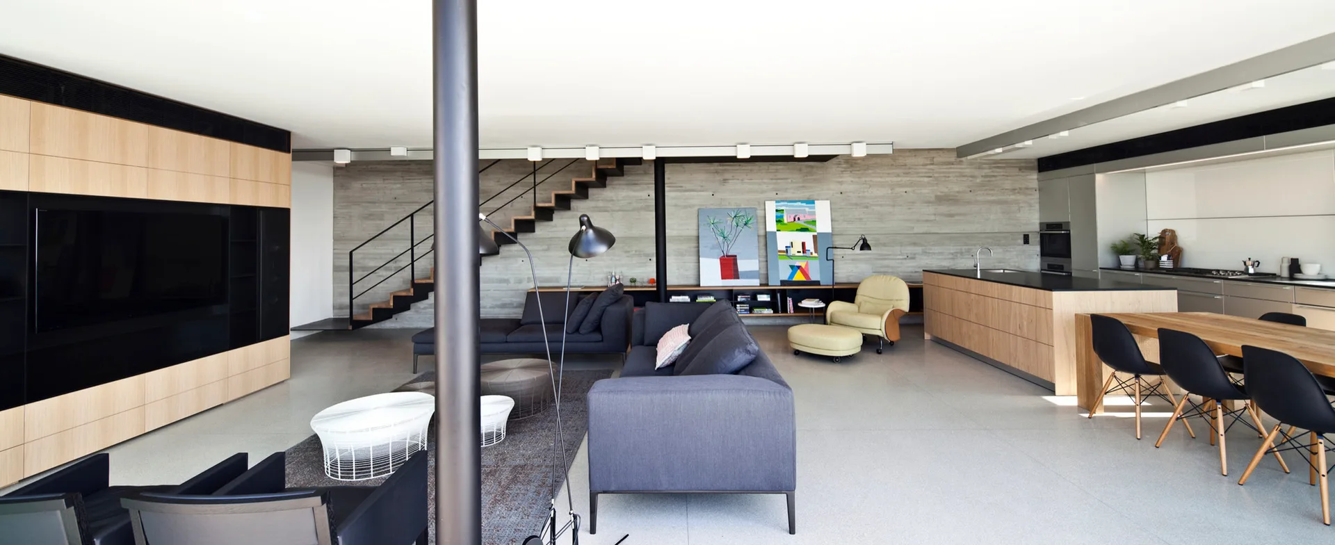 An open-concept living and dining area featuring a raw concrete wall, floating wooden stairs, dark grey sofas, and a wood-faced kitchen island.
