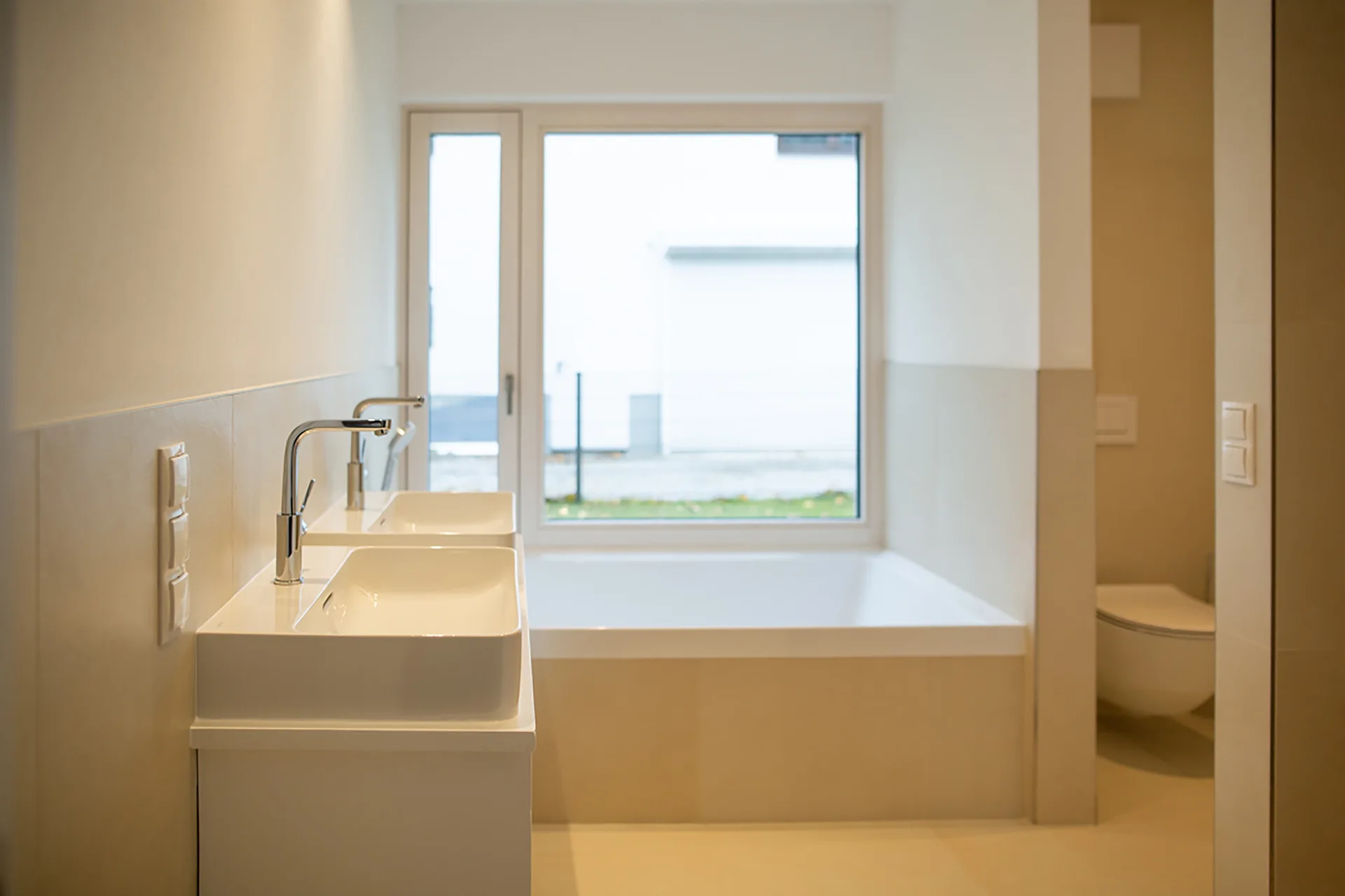 A modern beige bathroom featuring a white double sink vanity with chrome faucets, a built-in bathtub below a large window, and a wall-mounted toilet.