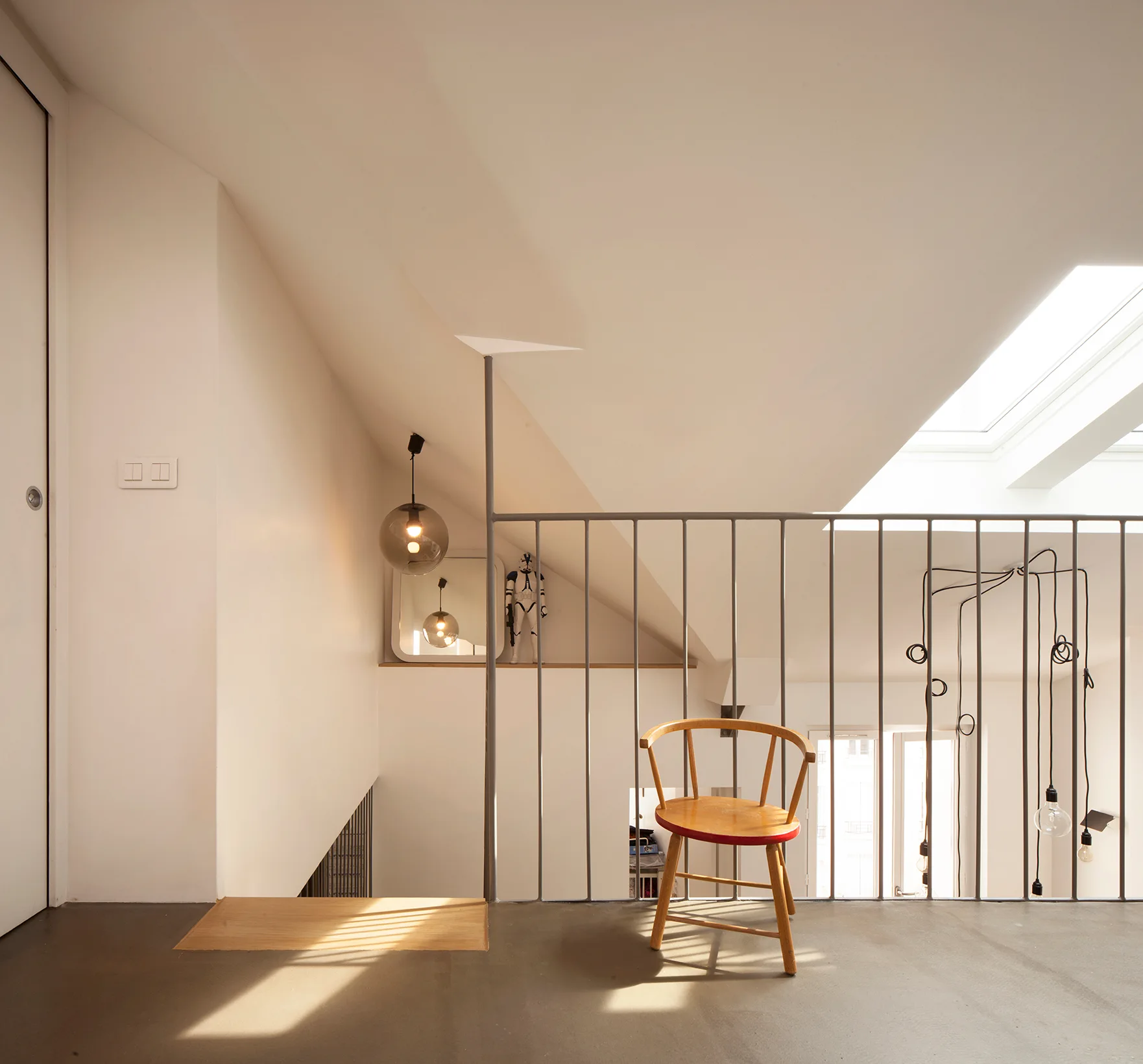 A minimalist loft landing features a single wooden chair next to a metal spindle railing under a sloped ceiling with a bright skylight.
