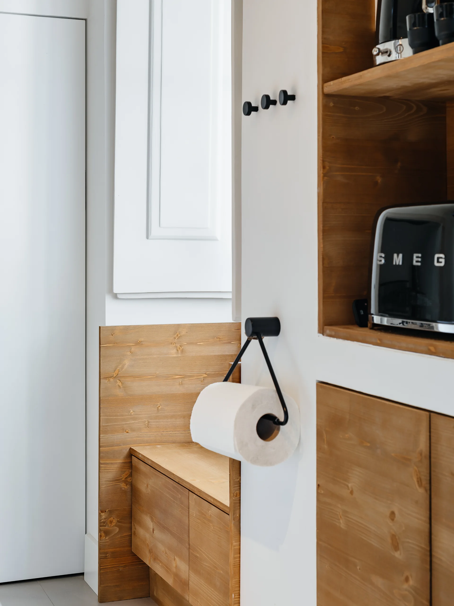 Built-in natural wood cabinetry features a black toaster on an open shelf alongside white walls fitted with black metal hooks and a roll holder.