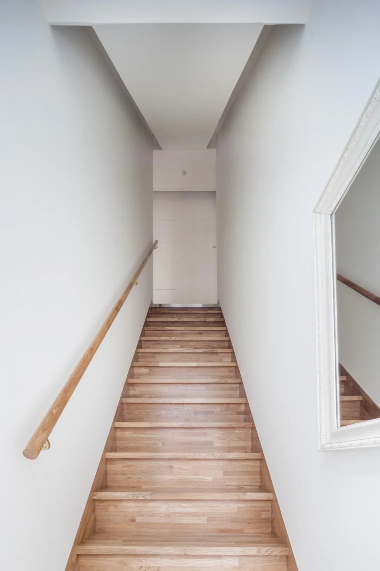 A narrow staircase featuring light wood treads, a matching wooden handrail, white walls, and a white framed mirror on the side.