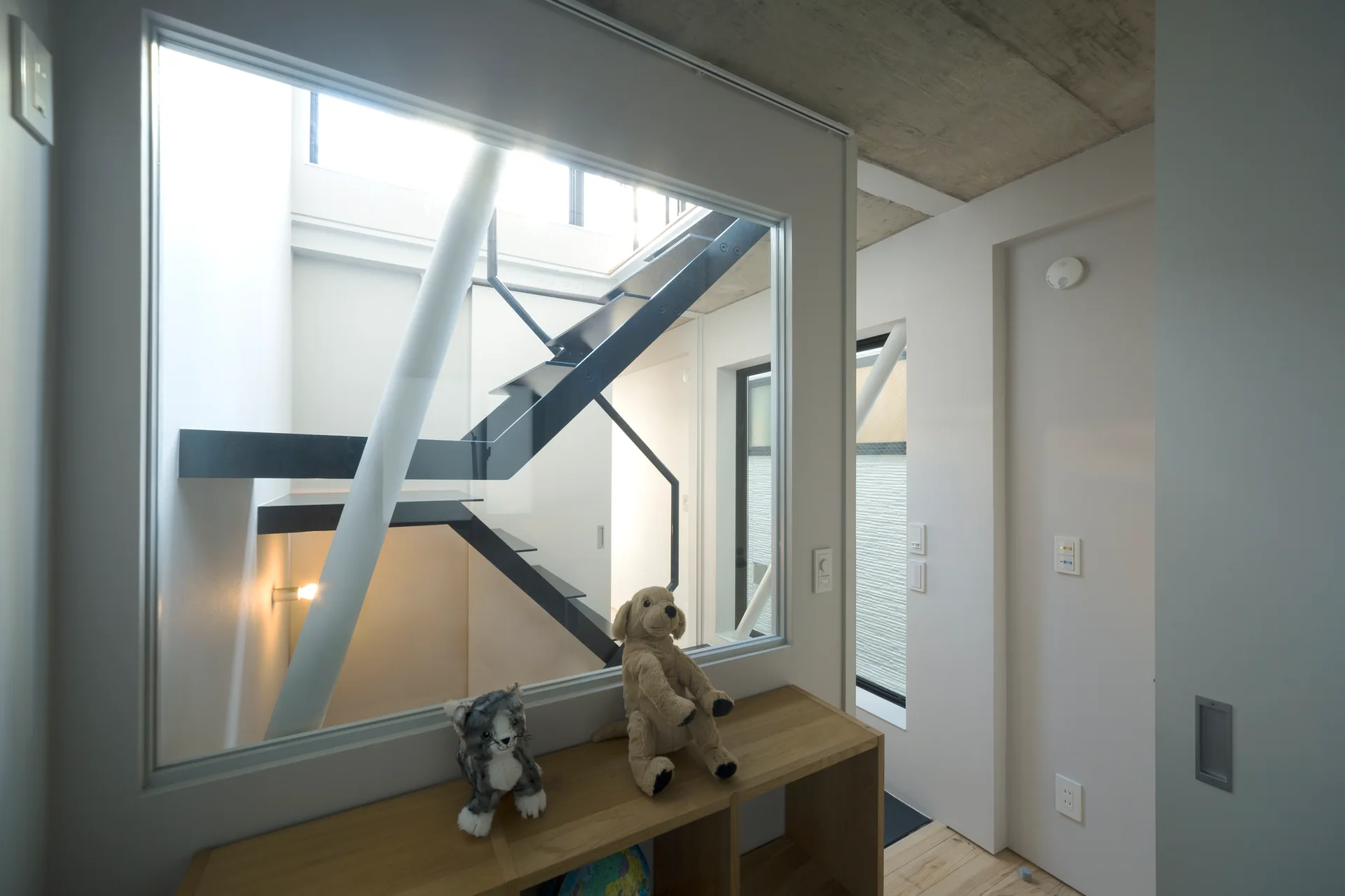 A light wood console table stands before a large interior glass window showcasing a modern black metal staircase and concrete ceiling.