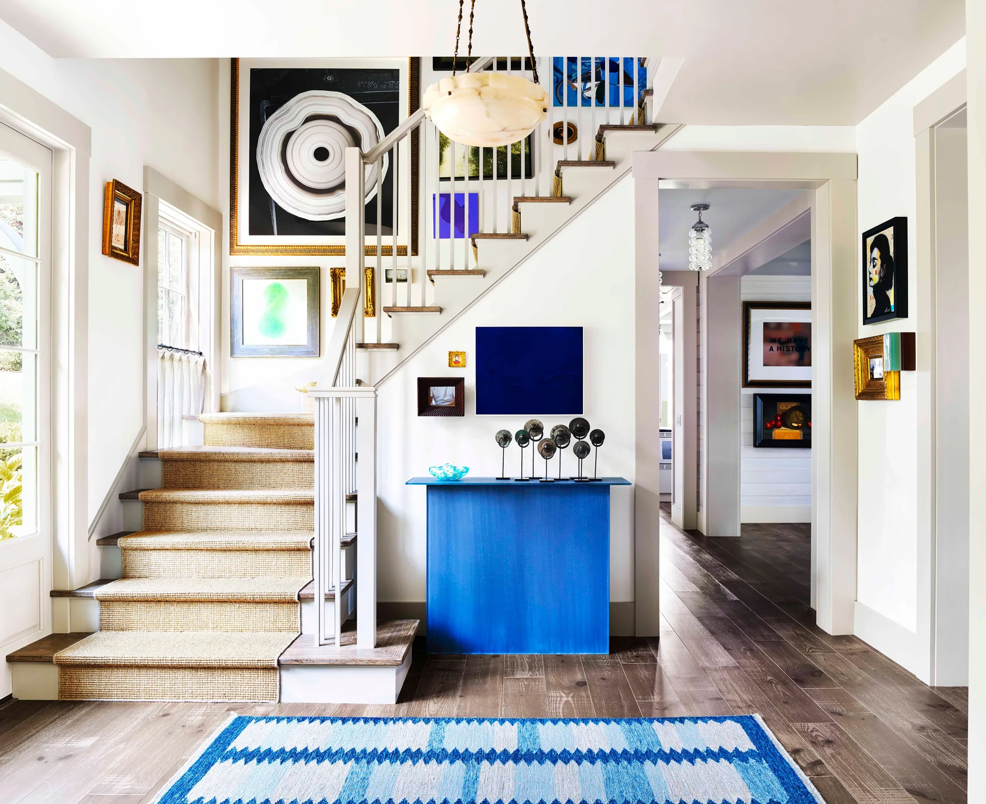 A light-filled foyer features a wood staircase with a woven runner, a bright blue console table, and an eclectic gallery wall.