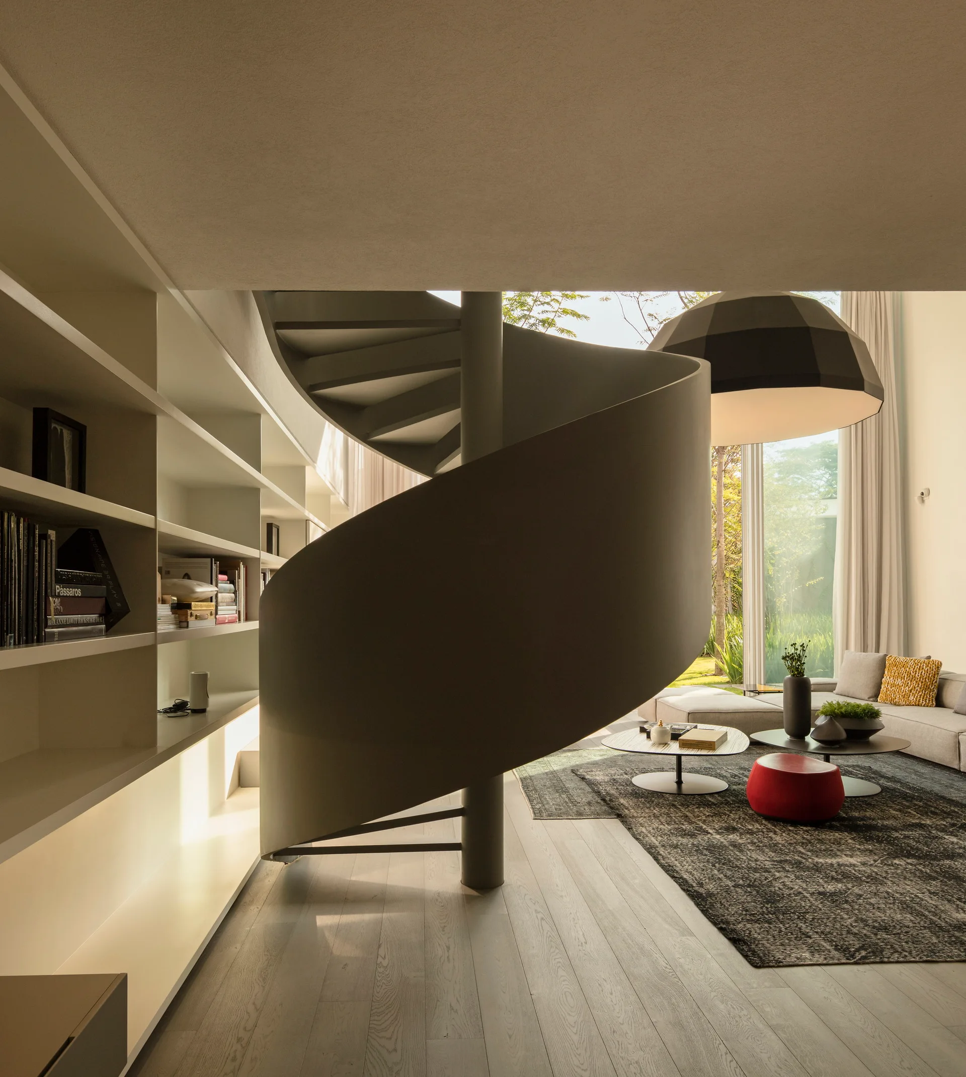 A modern living room features a dark sculptural spiral staircase next to built-in white bookshelves and a seating area with a sofa and red ottoman.