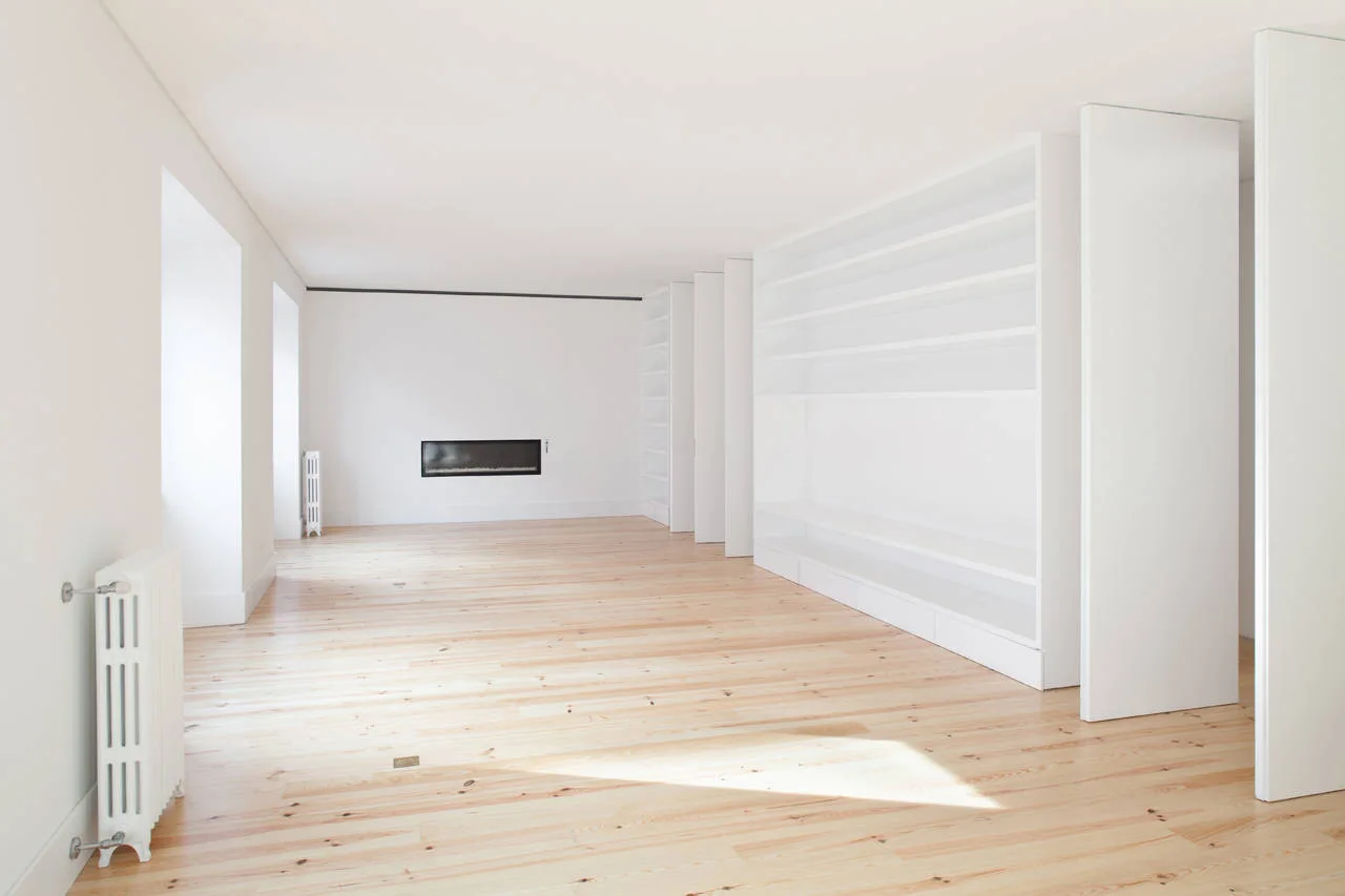 An empty minimalist room featuring light wood floors, matte white walls, a built-in shelving unit with sliding white panels, and a recessed fireplace.