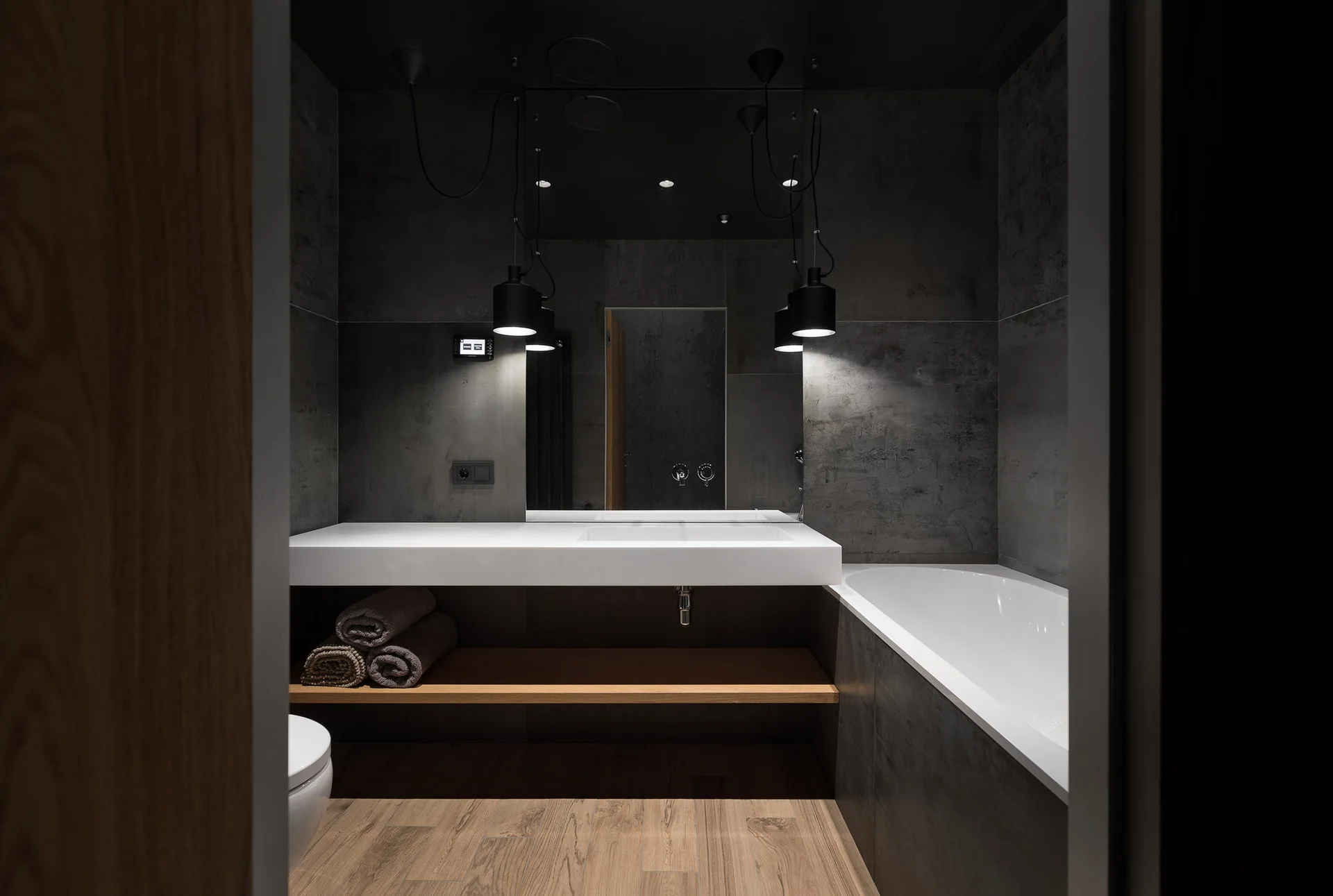 A modern bathroom with dark charcoal tiled walls, a white floating washbasin vanity, a light wood shelf, and black cylindrical pendant lights.