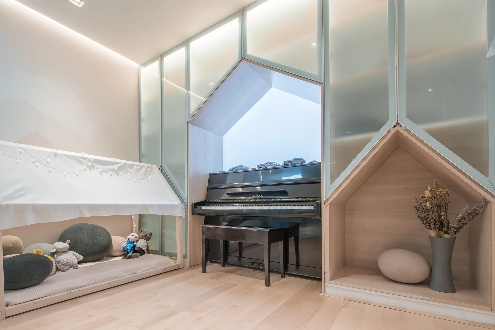 A children's playroom features a black piano nested in a house-shaped wooden niche alongside frosted glass cabinets and a cozy play tent.